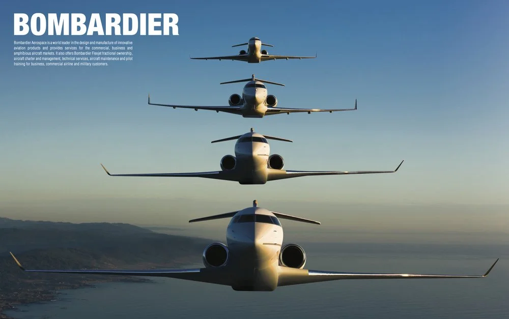 Five Bombardier airplanes flying in formation across a clear sky over a body of water with distant hills.