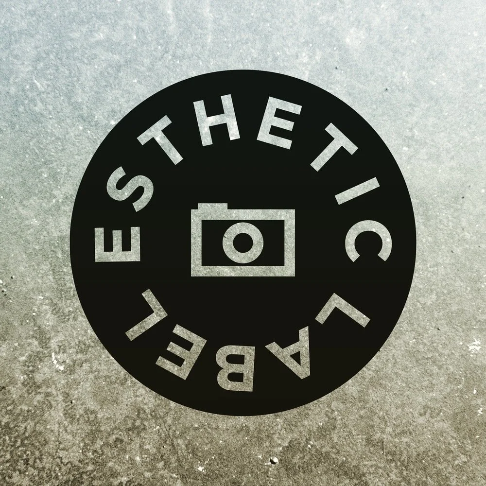 A circular black and white sticker on a textured concrete surface that reads 'BE ALERT THE TICKET' around a camera icon in the center.