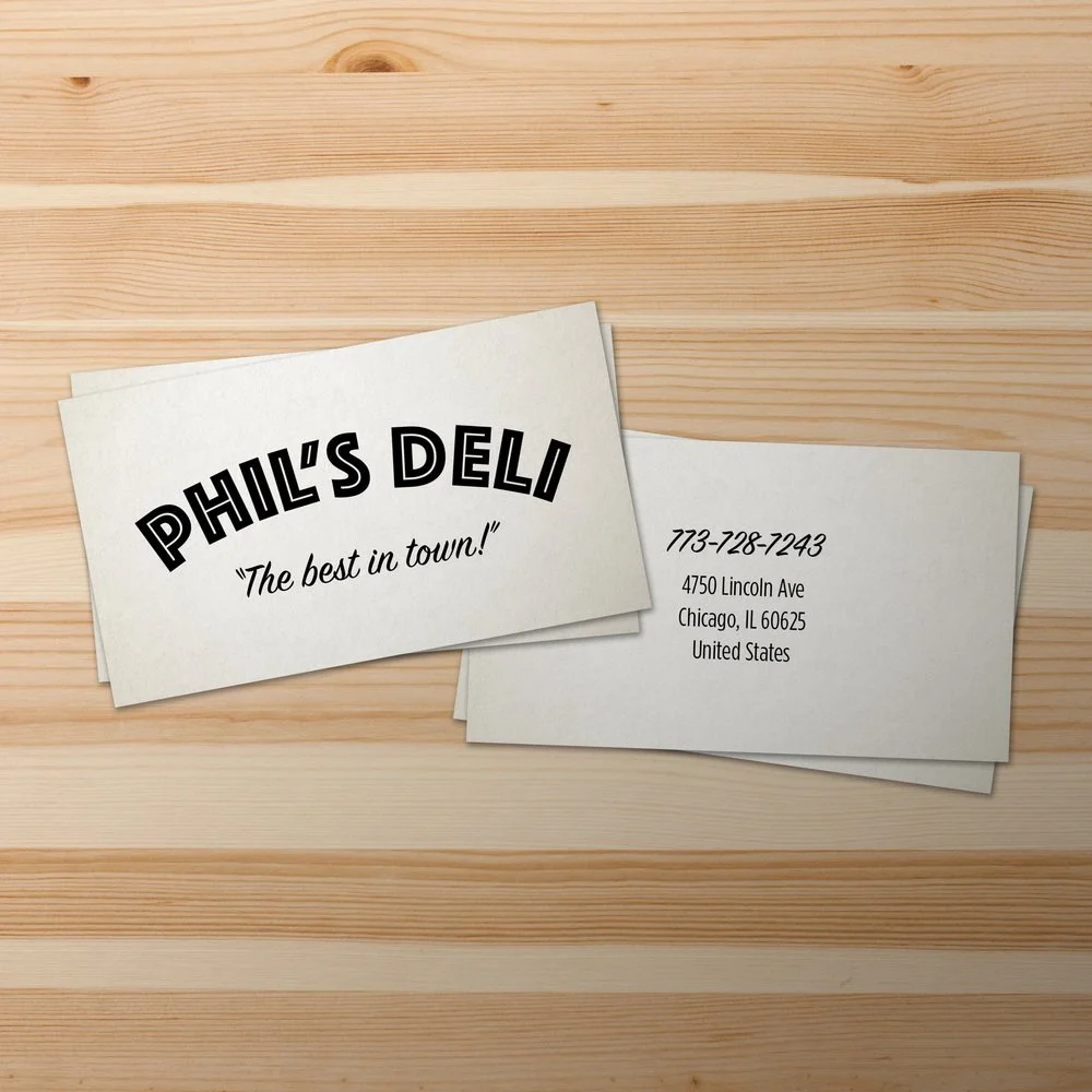 Business cards for Phil's Deli with contact details on a wooden surface.