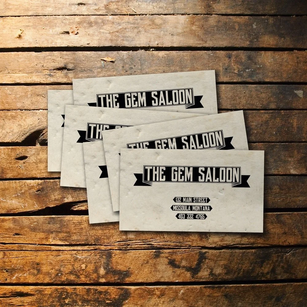 Four business cards for The Gem Saloon on a rustic wooden surface.