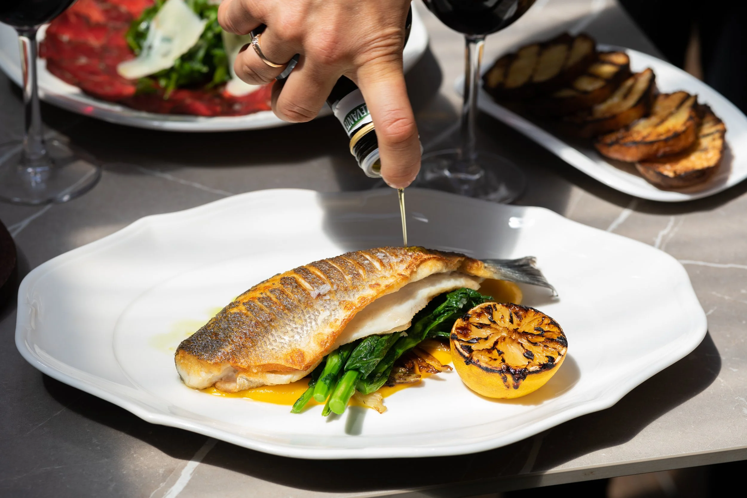 A plated dish featuring a grilled fish fillet topped with olive oil, served with grilled lemon, sautéed greens, and a yellow sauce. There are two glasses of red wine and additional dishes in the background.