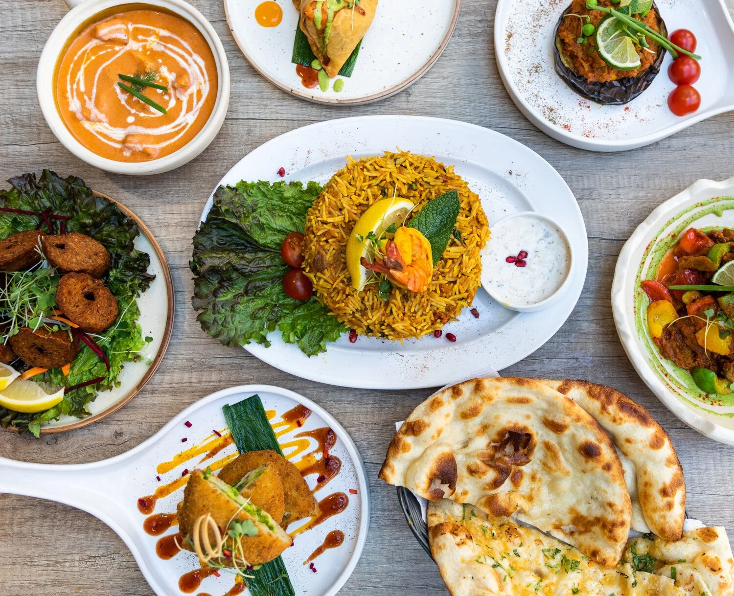 A variety of Indian dishes including naan bread, basmati rice, curry, grilled vegetables, fried snacks, and chutneys on different plates on a wooden table.