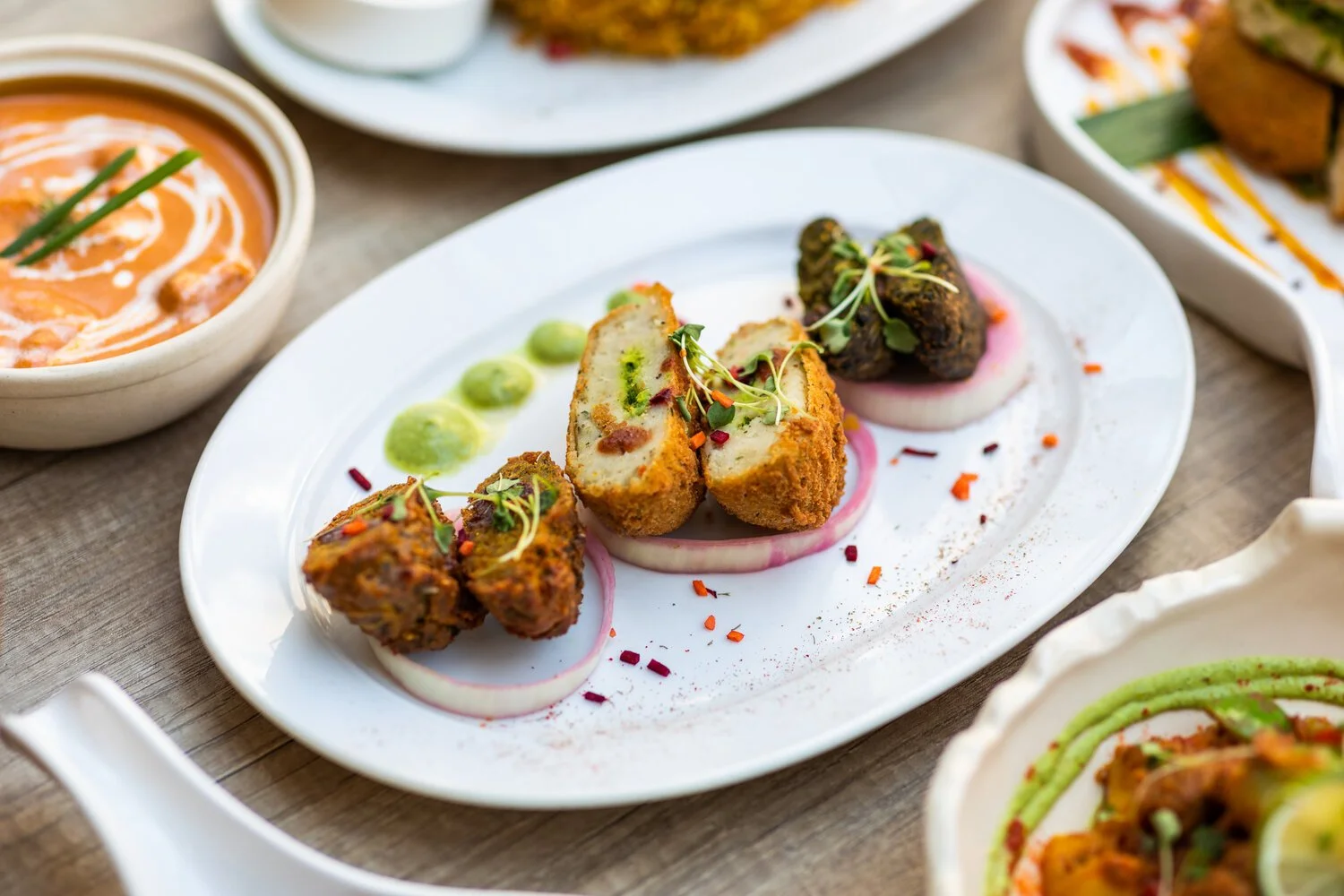 Plate with assorted Indian appetizers including fried stuffed balls garnished with microgreens, sliced onions, and green sauce, surrounded by bowls of curry and other Indian dishes.