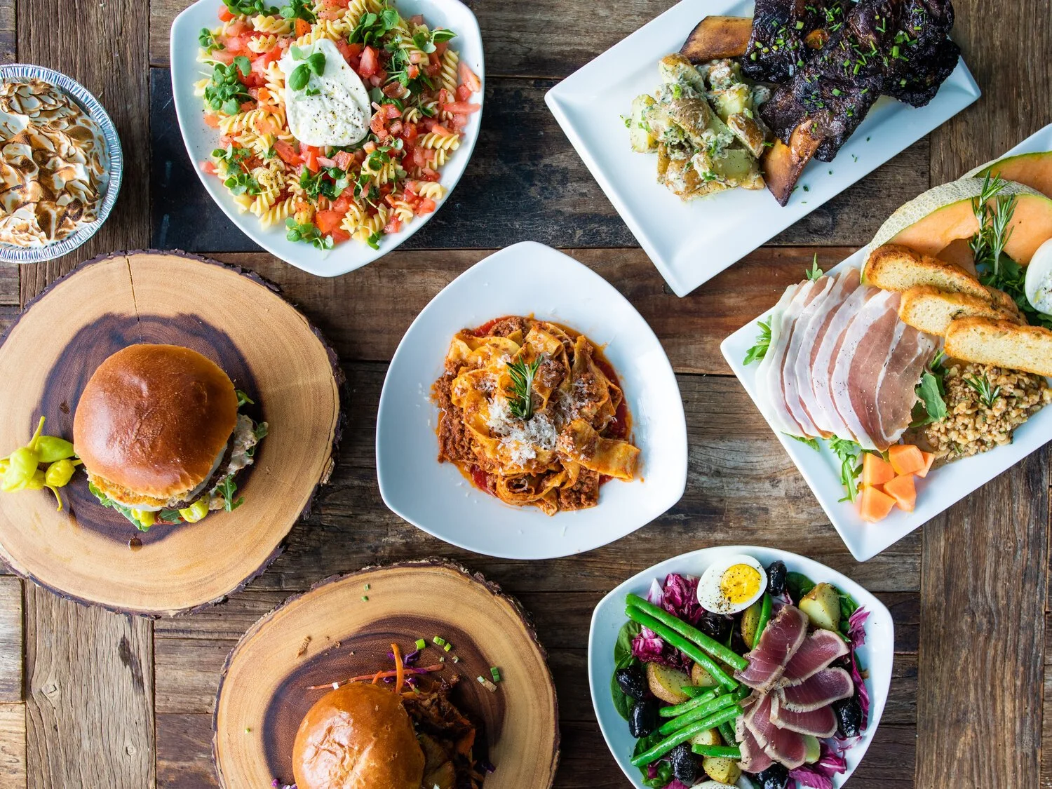 A rustic wooden table with various dishes, including pasta salad, roasted meat, a sandwich with prosciutto and grains, a burger on a wooden plate, a pasta dish, and a salad with greens and vegetables.