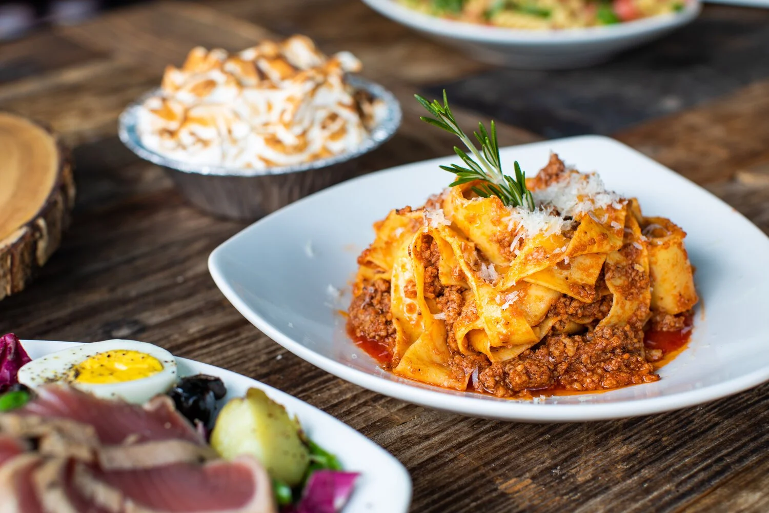 Bowl of pasta with ground meat and grated cheese, garnished with a sprig of rosemary, served on a white plate with other dishes visible in the background.