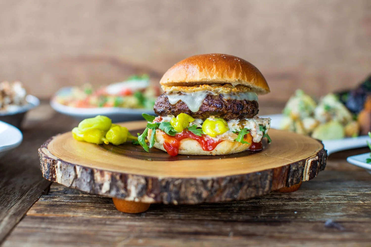Gourmet cheeseburger with toppings on a wooden serving board, with side dishes in the background.