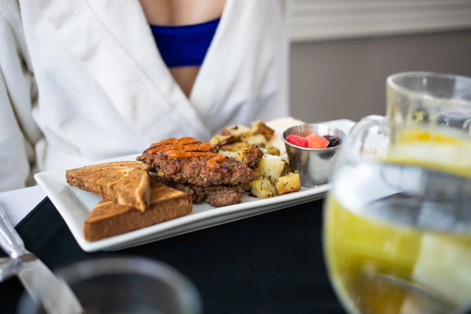 A breakfast plate with toast, a sausage patty, scrambled eggs with peppers, and a side of mixed berries, on a black table with a person's torso visible wearing a white shirt and blue top.