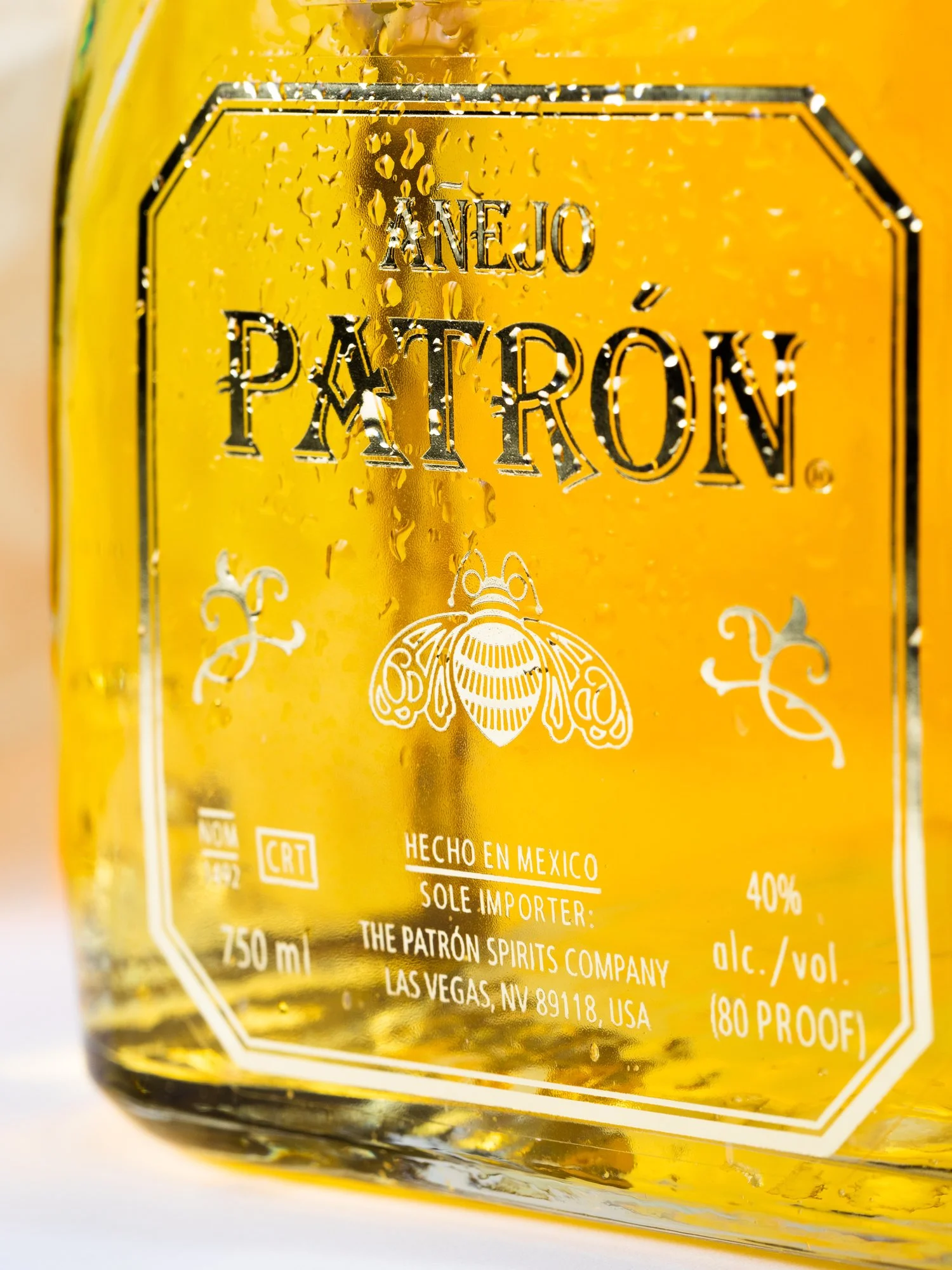 Close-up of a bottle of Anjeo Patrón tequila with condensation droplets on the surface, showing the label with gold color and black text, including a bee logo.