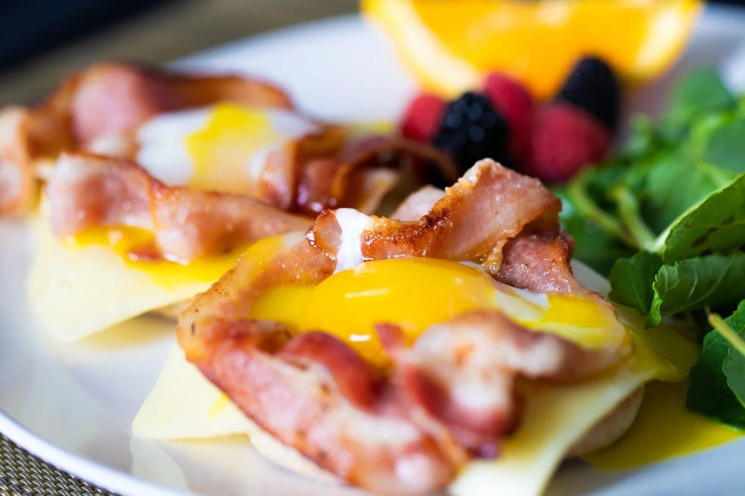 Close-up of two baked eggs with bacon on top, served on a white plate with mixed berries and leafy greens as garnish.