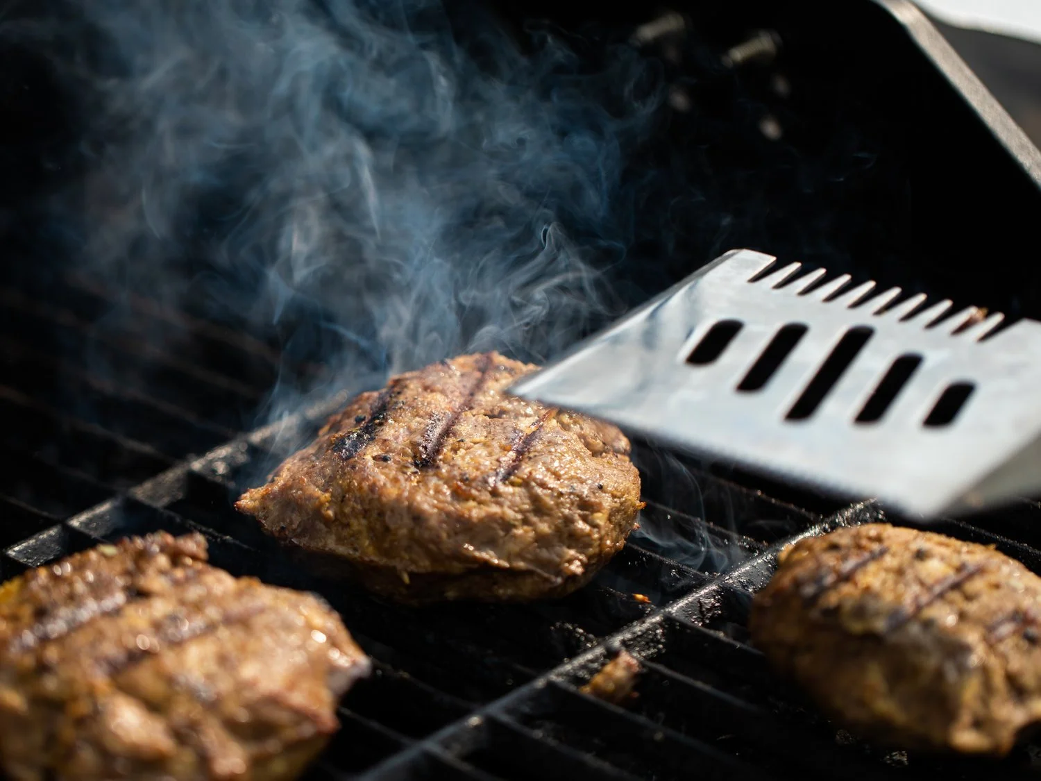 Three seasoned beef patties cooking on a grill with smoke, and a metal spatula nearby.