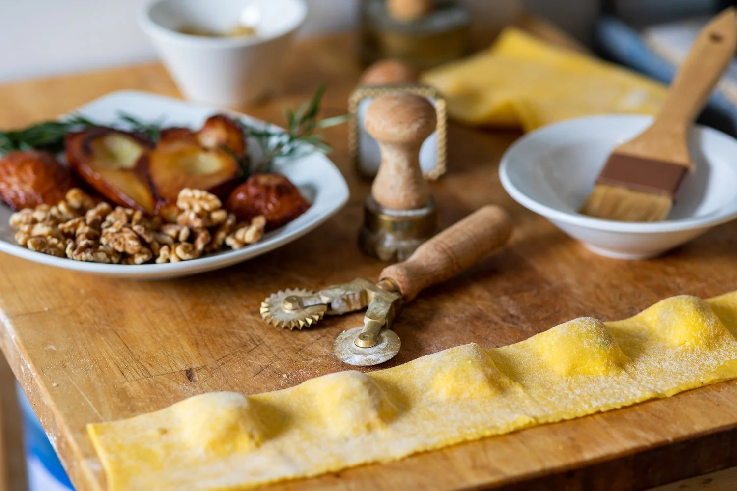 A wooden kitchen countertop with a plate of roasted peaches and walnuts, a bowl with a pastry brush, a wooden pepper grinder, a pasta cutter, and a sheet of uncooked yellow pasta dough in the foreground.