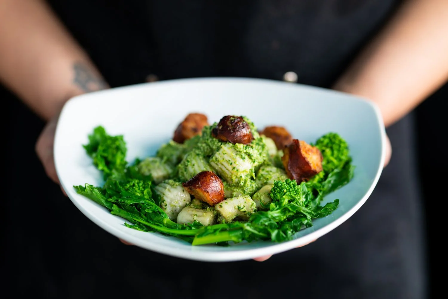 A white bowl of gnocchi with green pesto sauce, topped with crispy browned pieces, surrounded by fresh green parsley on a person's hands, against a dark background.