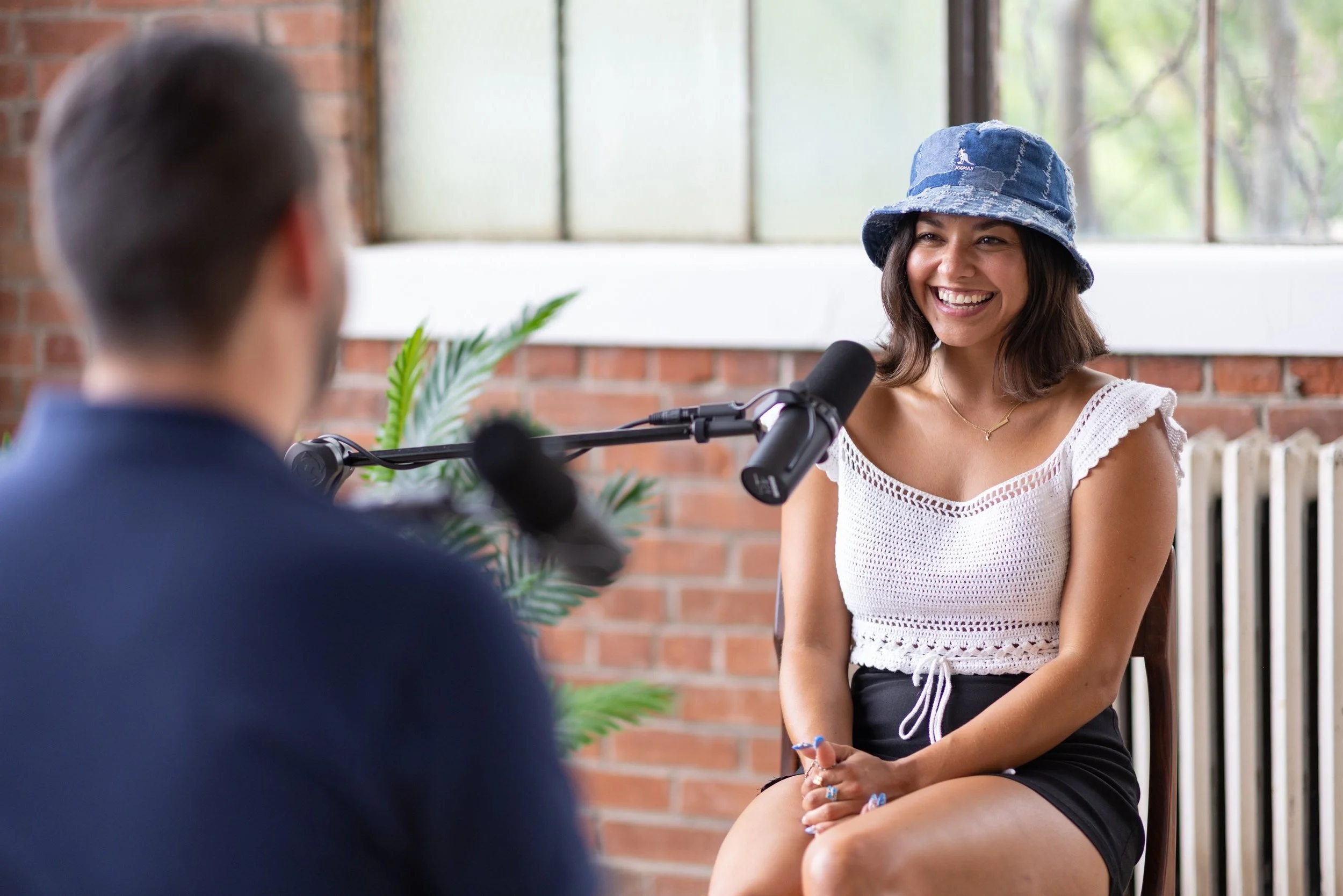 A woman wearing a blue bucket hat and white crochet top sits in front of a microphone, smiling during an interview with a blurred man in the foreground against a brick wall background.