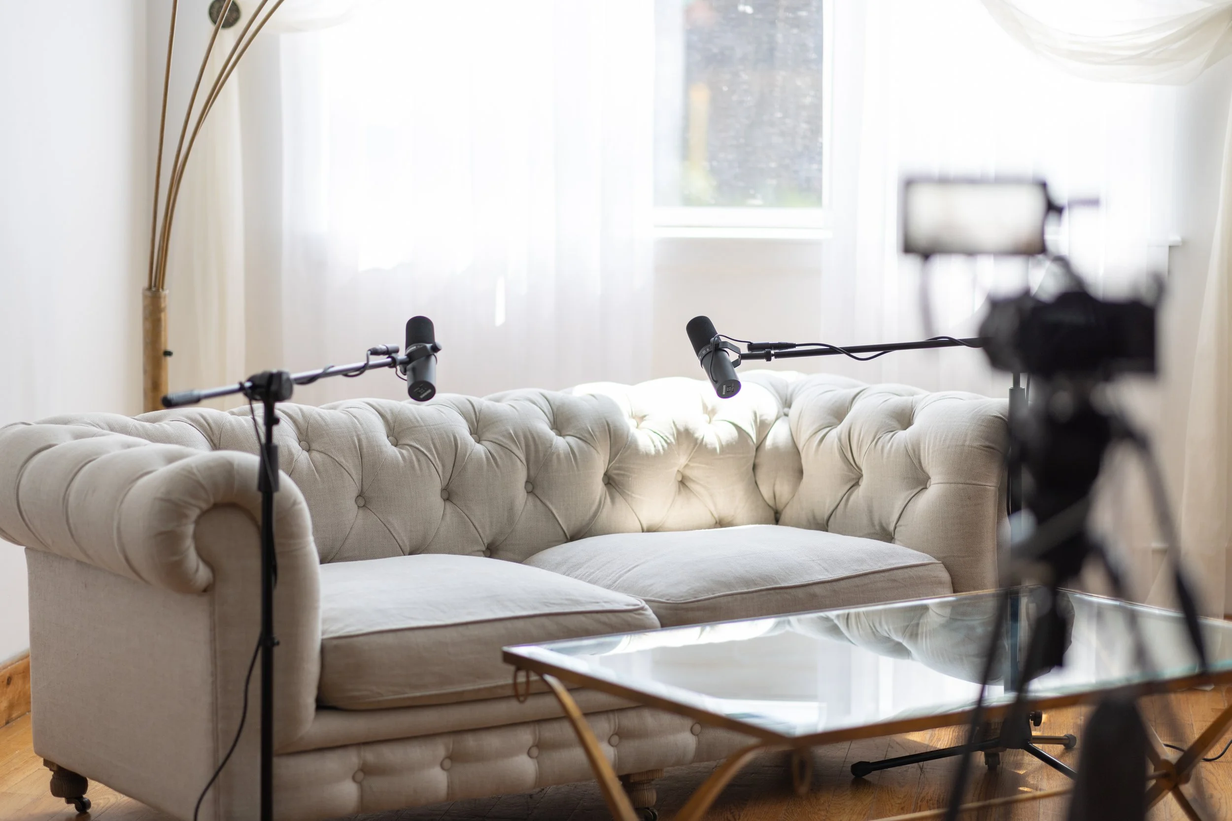 Living room scene with a beige tufted sofa, two microphones on stands, a camera on tripod, a glass coffee table, and a window with sheer curtains.
