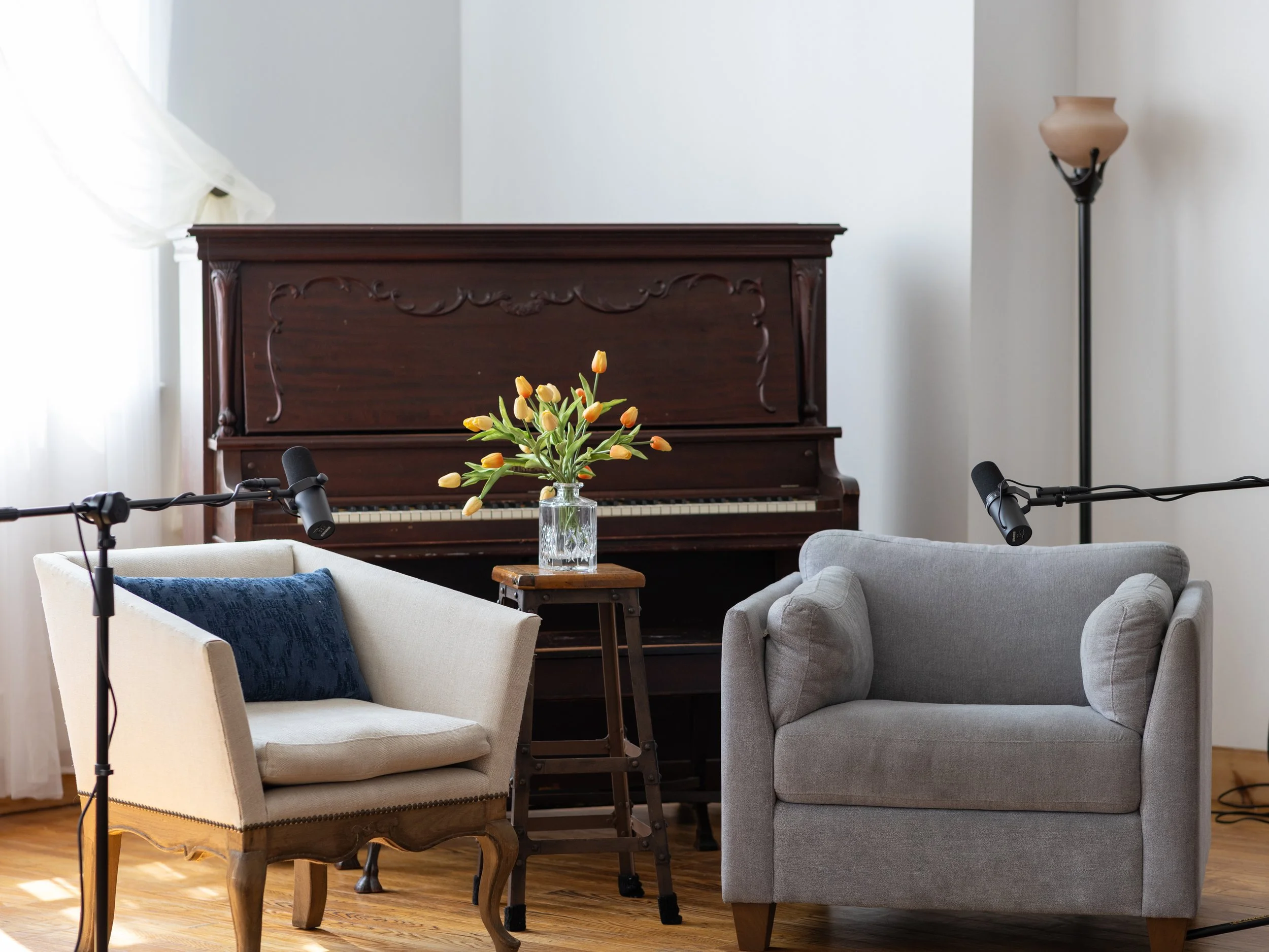 Interior of a cozy room with a wooden piano, two upholstered chairs, a small table with a vase of yellow tulips, and two microphones set up for recording or podcasting.
