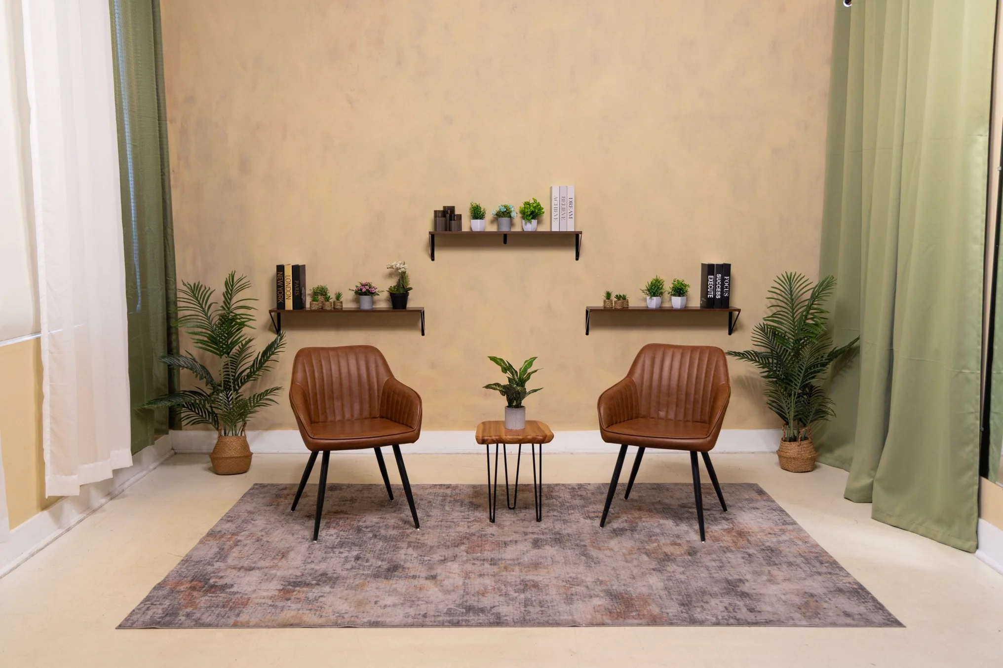A cozy sitting area with two brown leather chairs, a small wooden table with a potted plant, two green potted plants in woven baskets, three wall-mounted shelves with potted plants and books on beige walls, a patterned area rug, and curtains on either side.