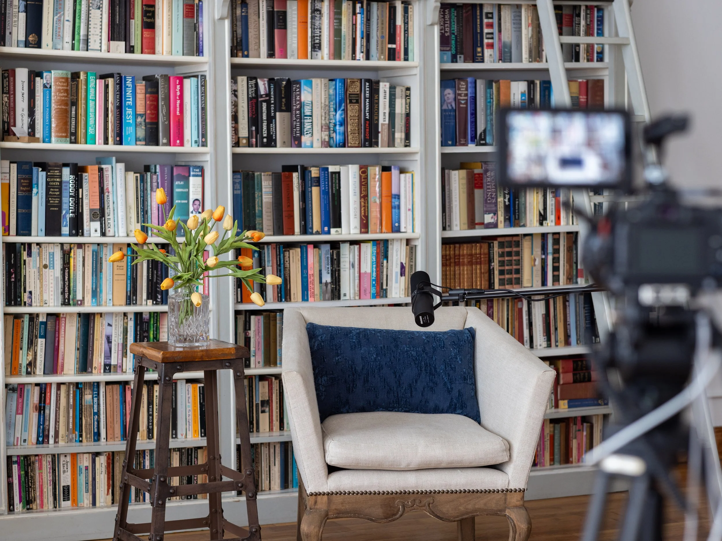 A cozy reading nook with a white armchair, a dark blue cushions, a side table with a glass vase filled with yellow tulips, and tall bookshelves filled with books in the background. A professional video camera on a tripod is visible in the foreground, and a microphone is mounted on the armchair.