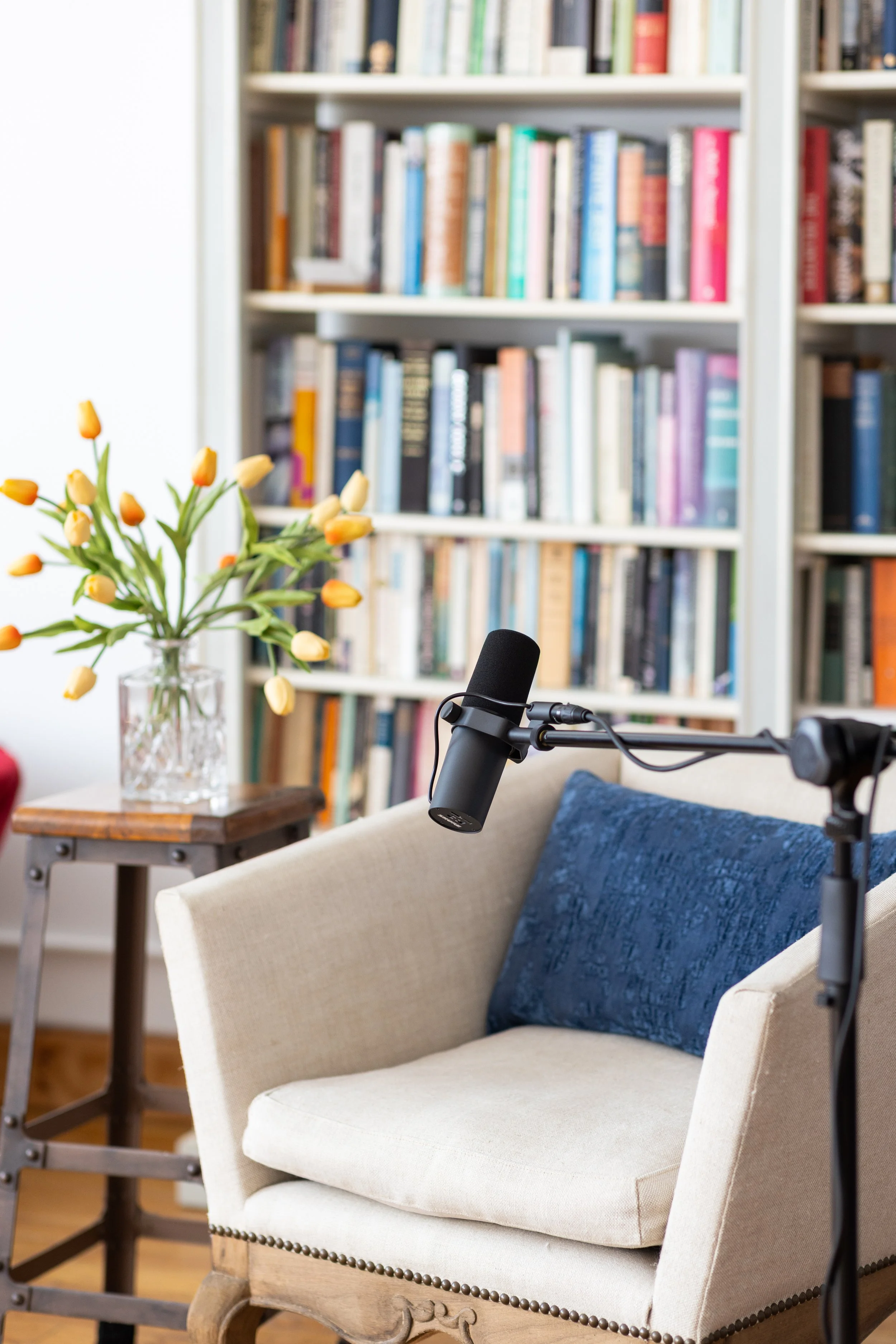 A cozy living room with a beige armchair, a blue cushion, a microphone on a stand, a wooden side table with a glass vase of yellow tulips, and a bookshelf filled with books in the background.