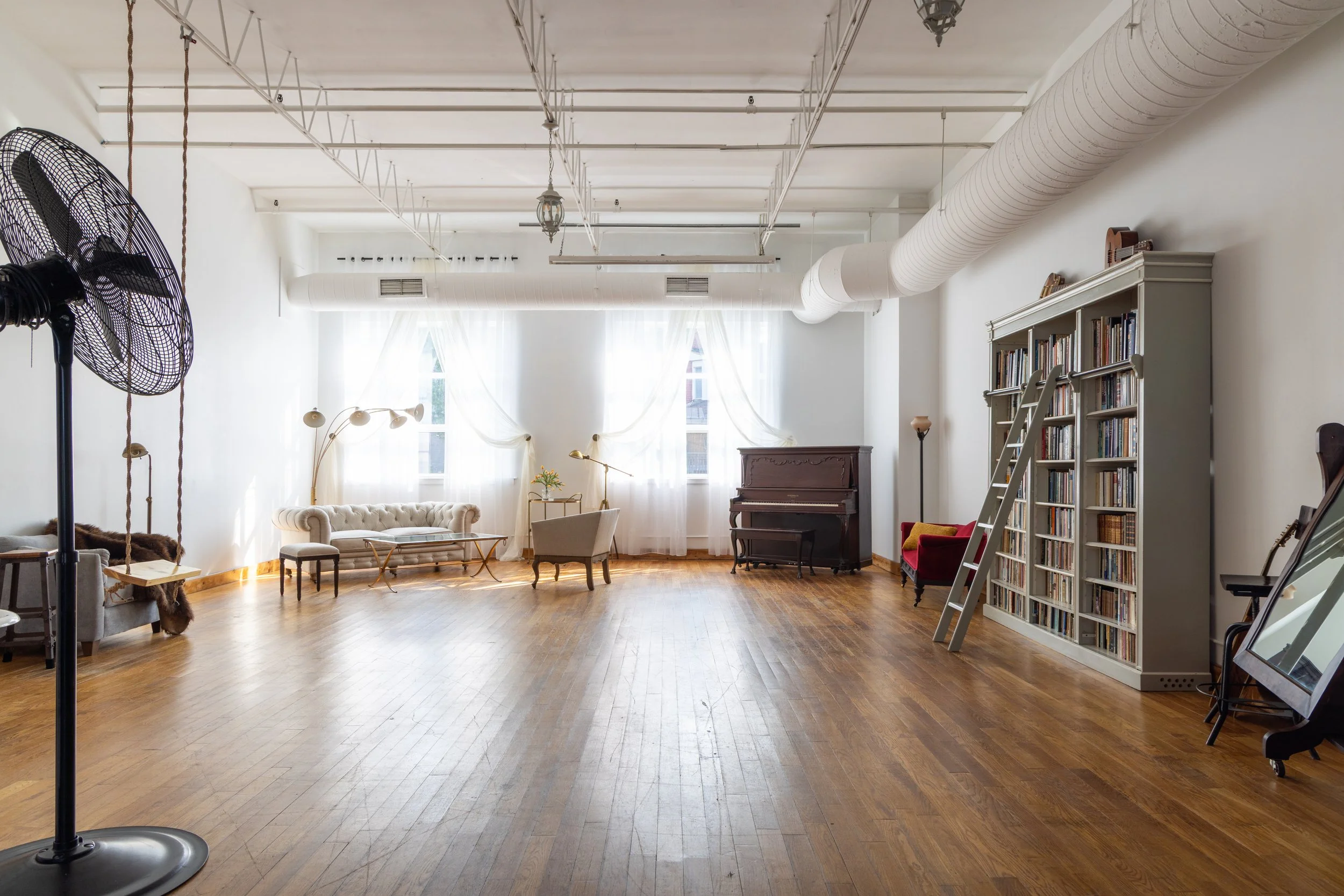 Spacious room with wooden floor, white walls, large windows with sheer curtains, and various vintage furniture including a piano, bookshelves, sofas, armchairs, and standing lamps.