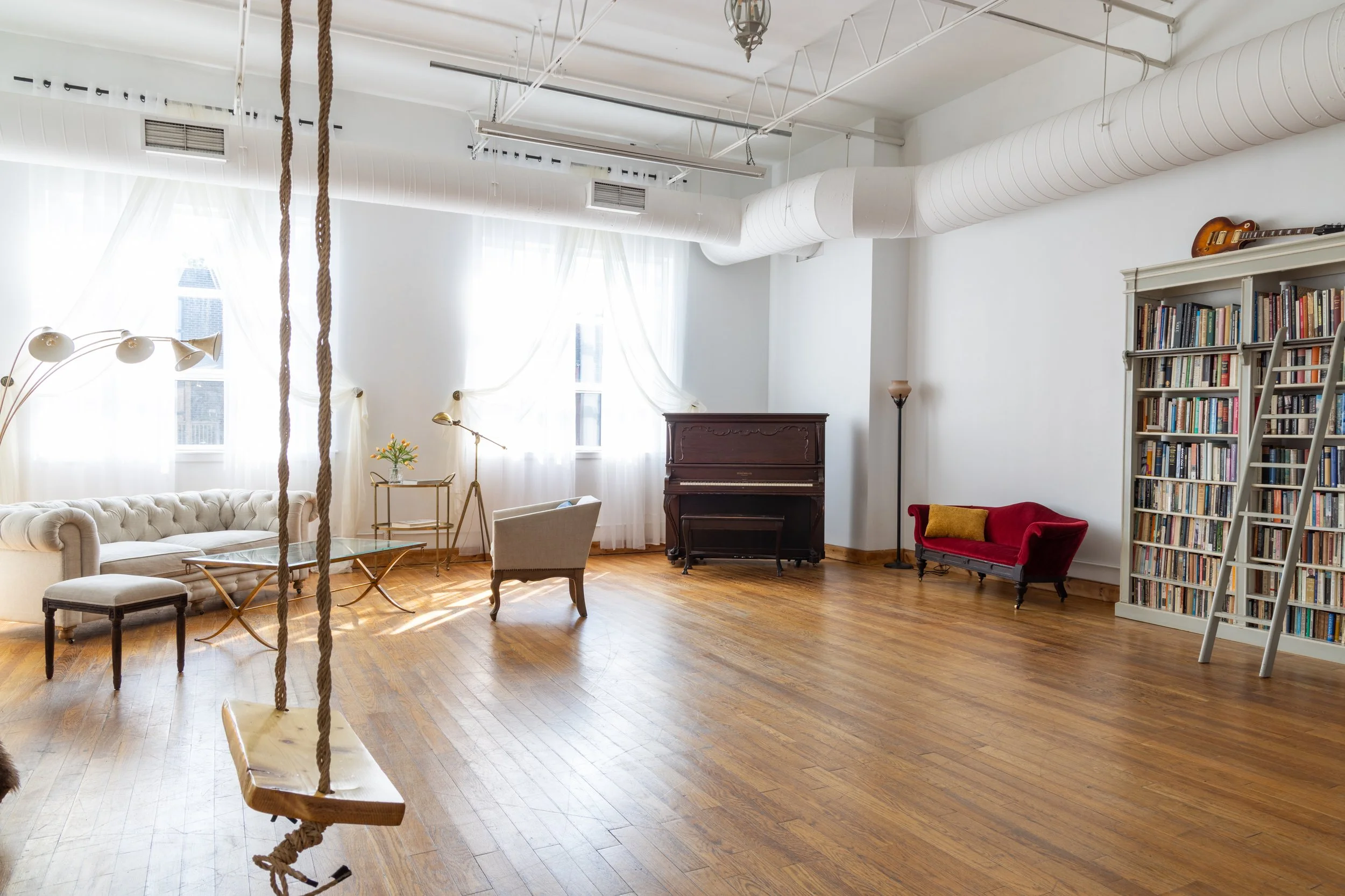 Bright living room with hardwood floors, white walls, large windows with sheer curtains, a beige sofa, a white armchair, a glass coffee table, a standing lamp, a wooden bookshelf filled with books, a ladder, a red velvet loveseat, an upright piano, and a wooden swing hanging from the ceiling.