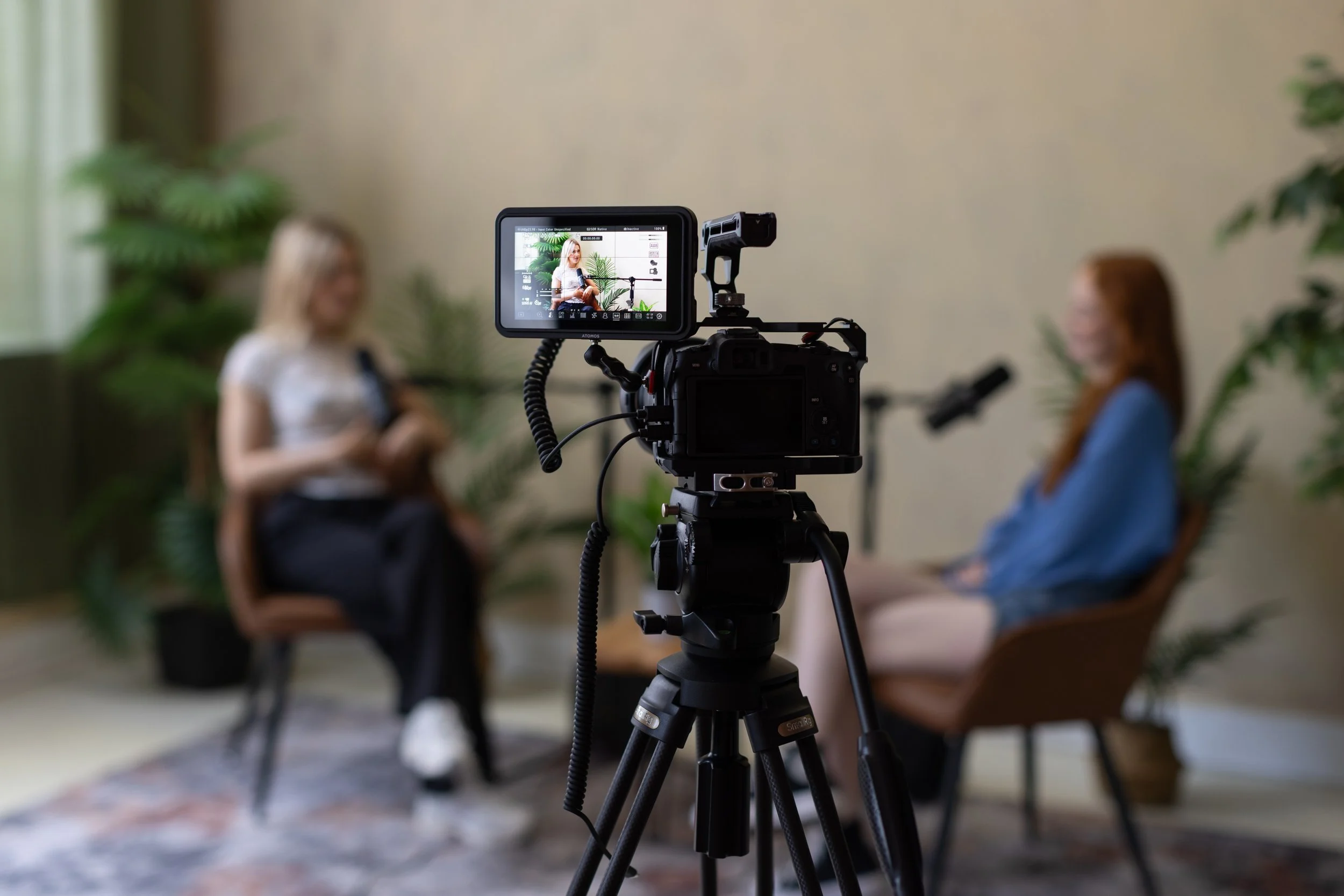 Two women sitting in chairs on a set with potted plants, being filmed with a camera on a tripod.