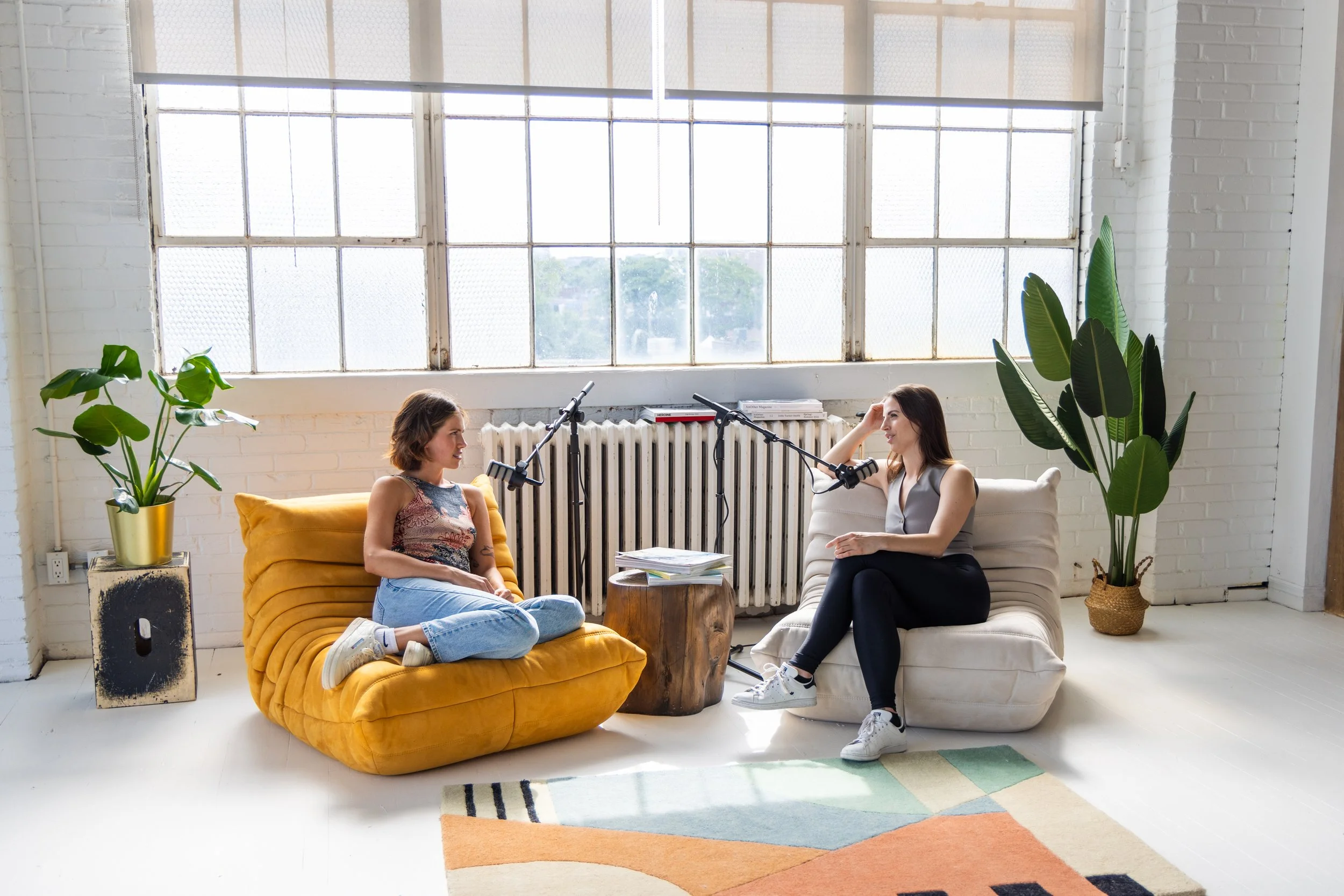 Two women sitting on couches having a conversation, surrounded by microphones, in a bright studio with large windows, potted plants, and a colorful rug.