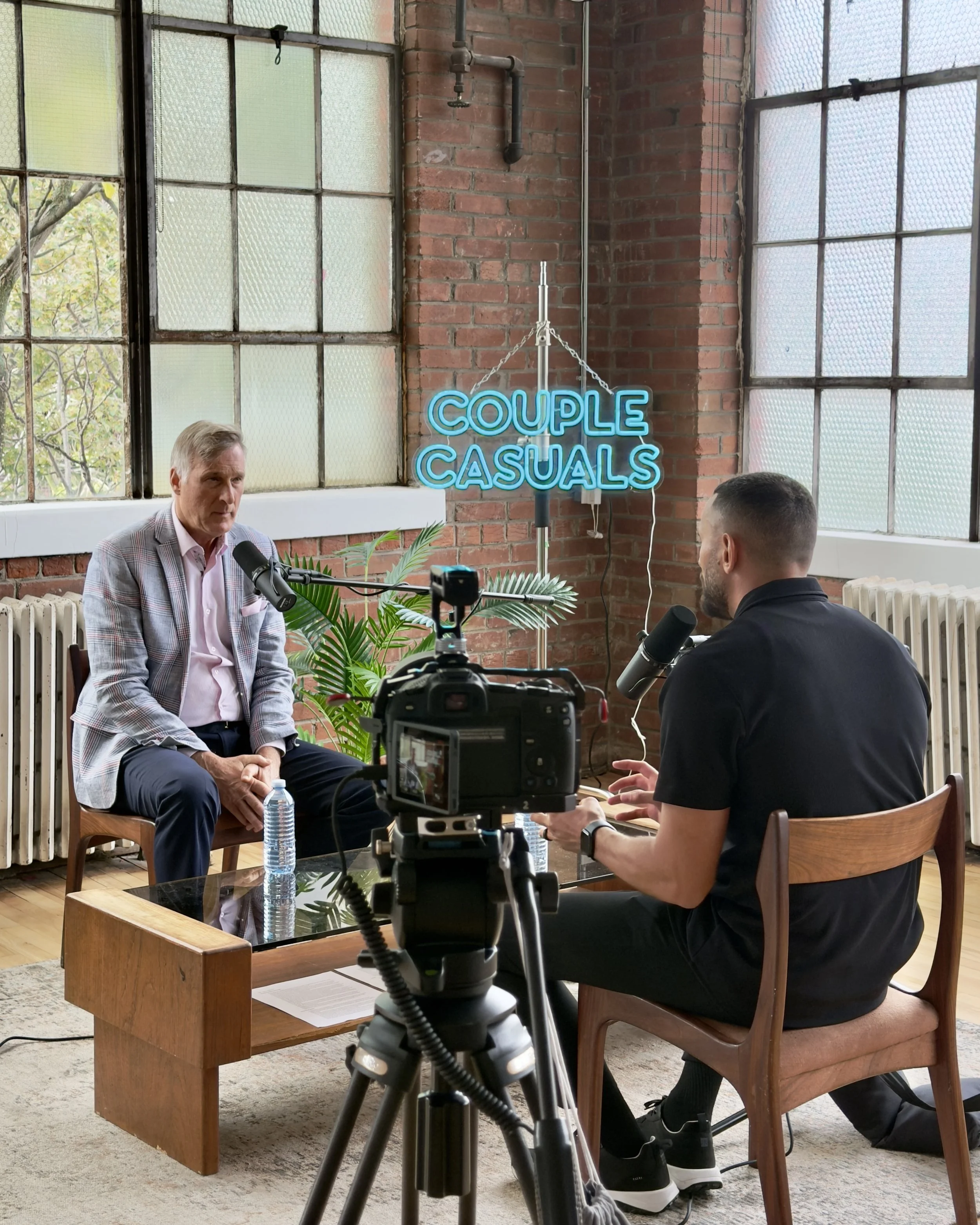 Two men having a conversation in an indoor studio with large windows and a brick wall, one wearing a light blazer and the other in black, with a camera recording them and a neon sign reading 'COUPLE CASUALS' in the background.