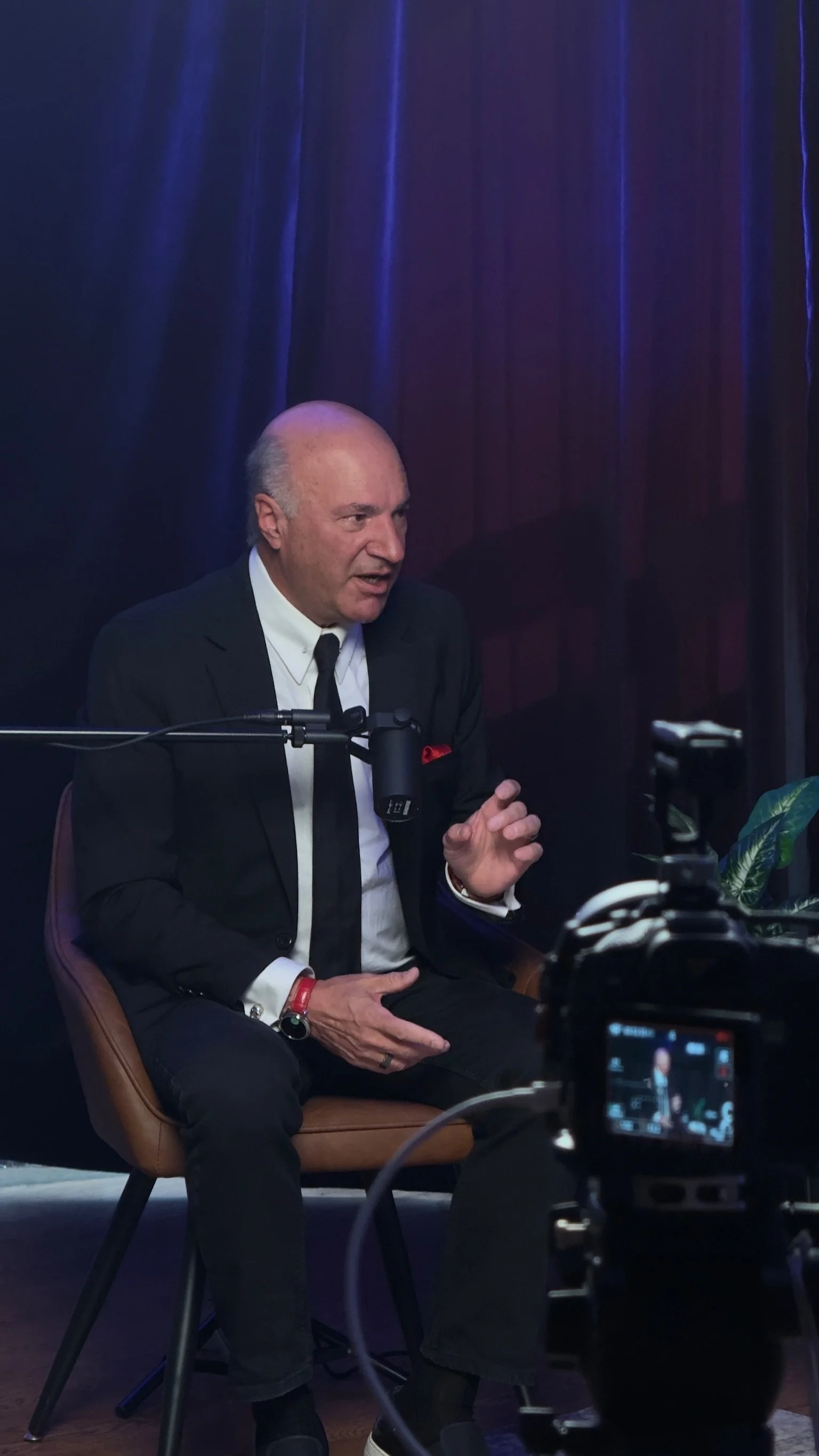 A man seated on a brown chair, dressed in a black suit with a white shirt and black tie, speaking during an interview or recording session, with a purple curtain backdrop and camera equipment visible.