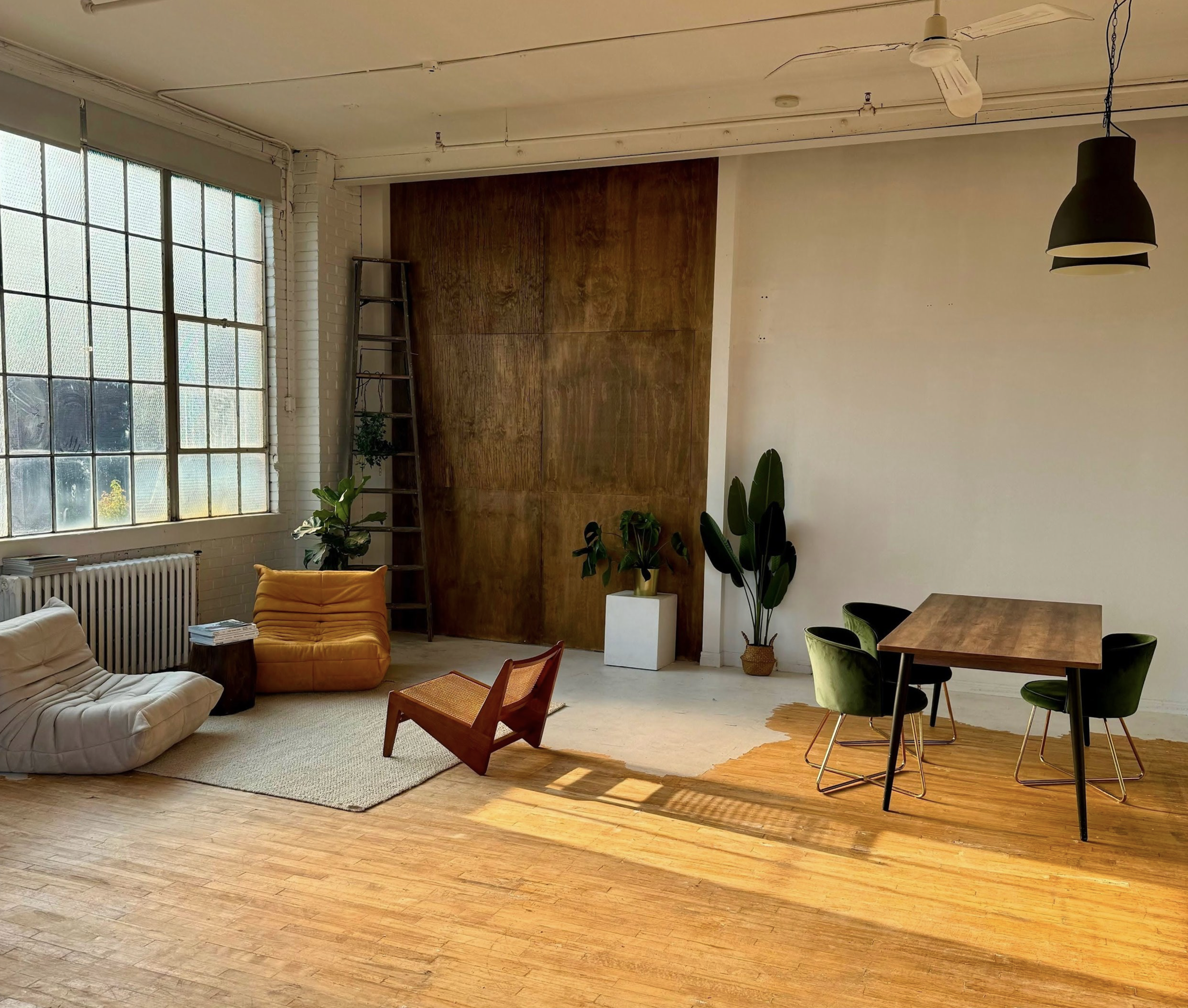 Living space with a large window on the left, furnished with modern chairs, a wooden table with four green chairs, plants, and a ladder against a wooden accent wall.