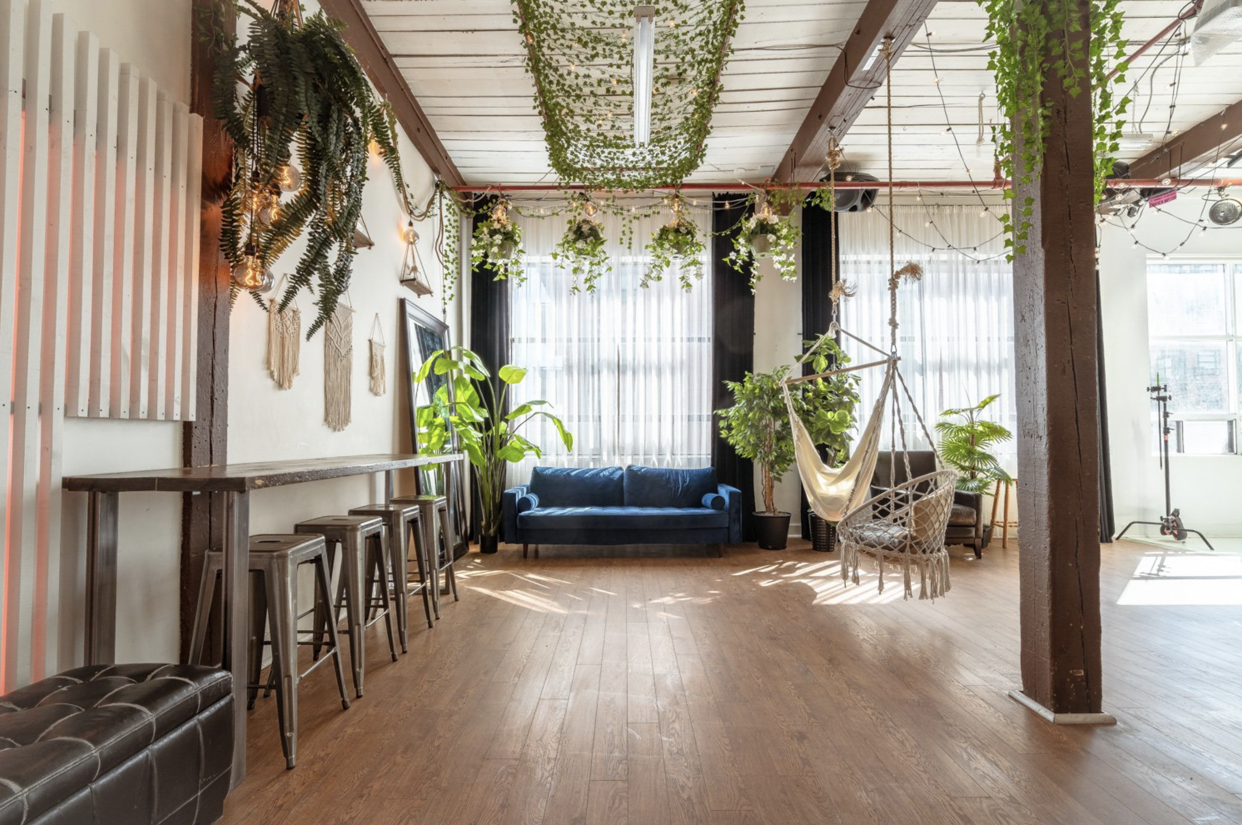 Bright, cozy lounge area with a blue velvet sofa, hanging chair, and potted plants. Decor includes string lights, hanging greenery, and wall art, with large windows allowing natural light.