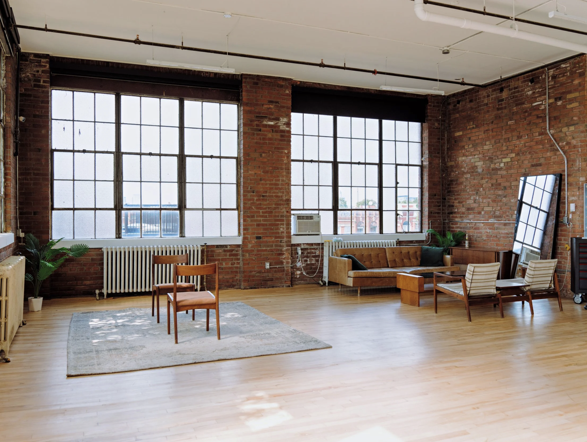 Spacious loft with large industrial windows, exposed brick walls, hardwood floors, and vintage furniture including a sofa, chairs, and mirror.