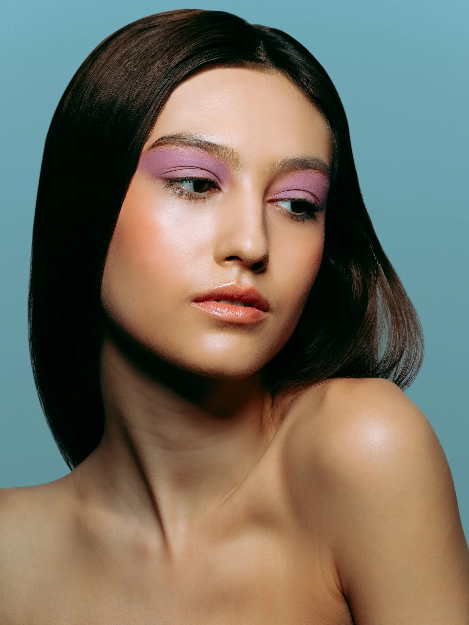 Close-up of a woman with dark brown hair and pink eyeshadow, looking to the side with a neutral expression against a light blue background.