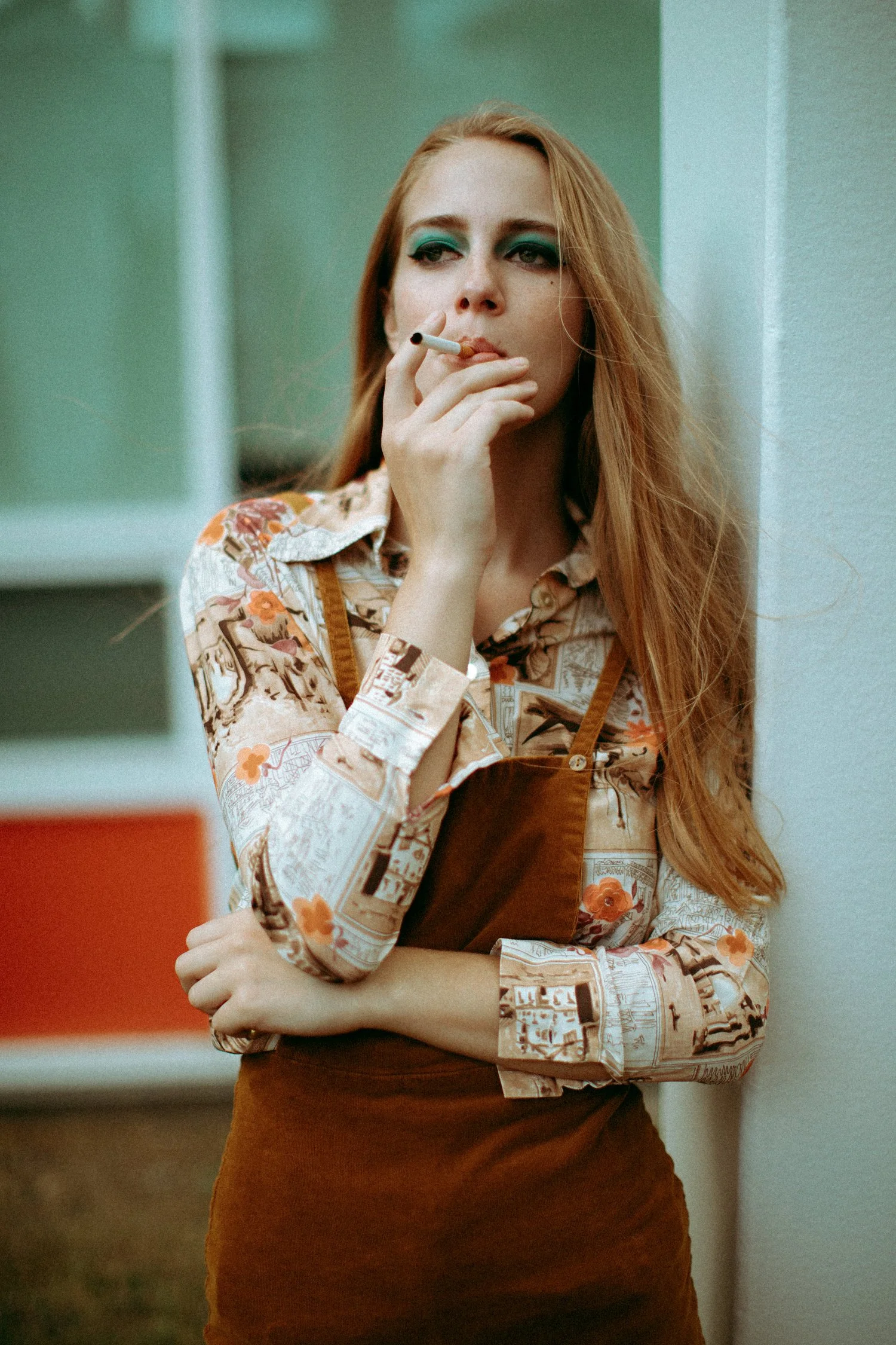 A woman with long red hair and teal eyeshadow is smoking a cigarette, standing against a wall.