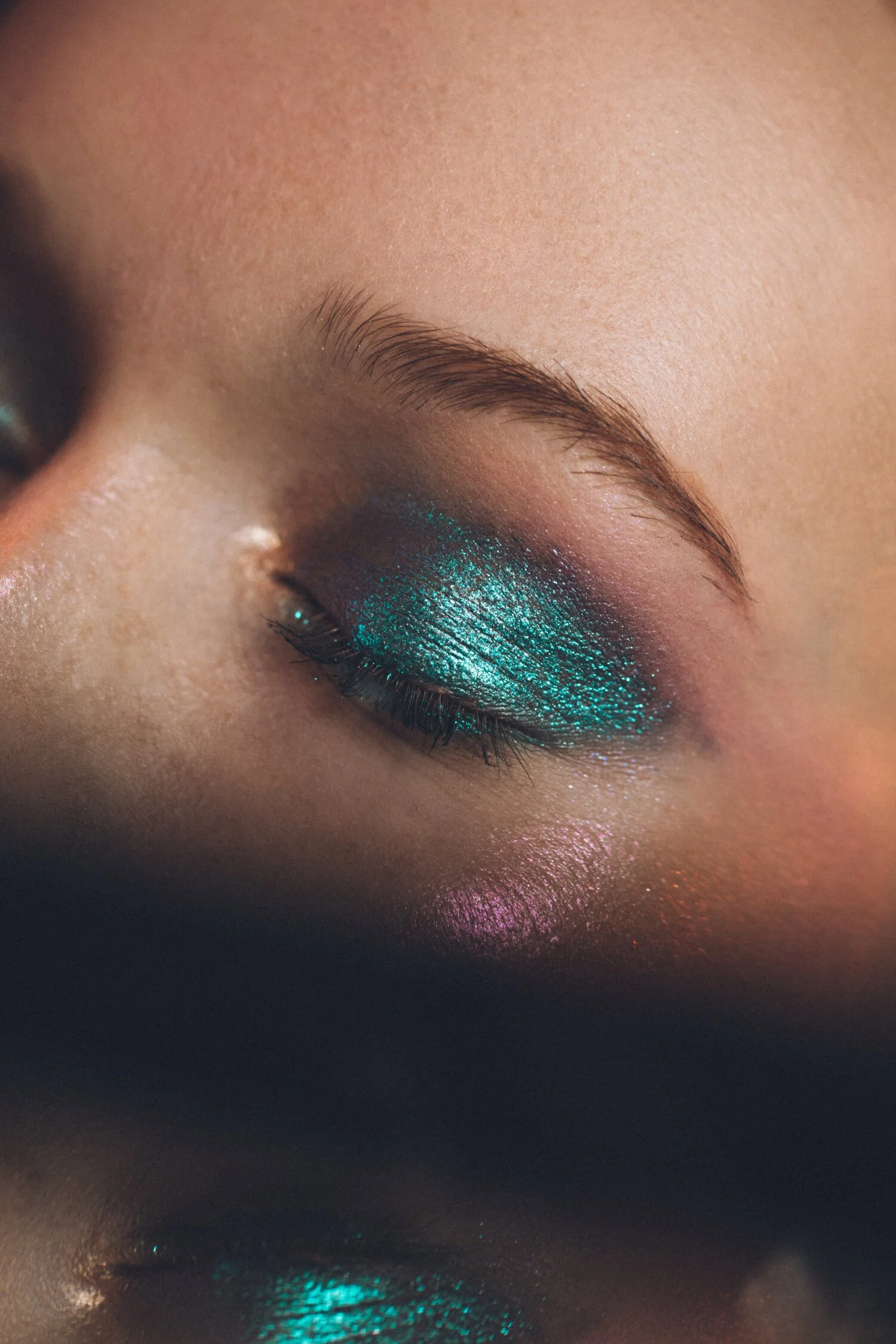 Close-up of a person's eye with vibrant turquoise and purple eyeshadow and shimmering makeup, looking downward with a reflection of the eye and makeup visible below.
