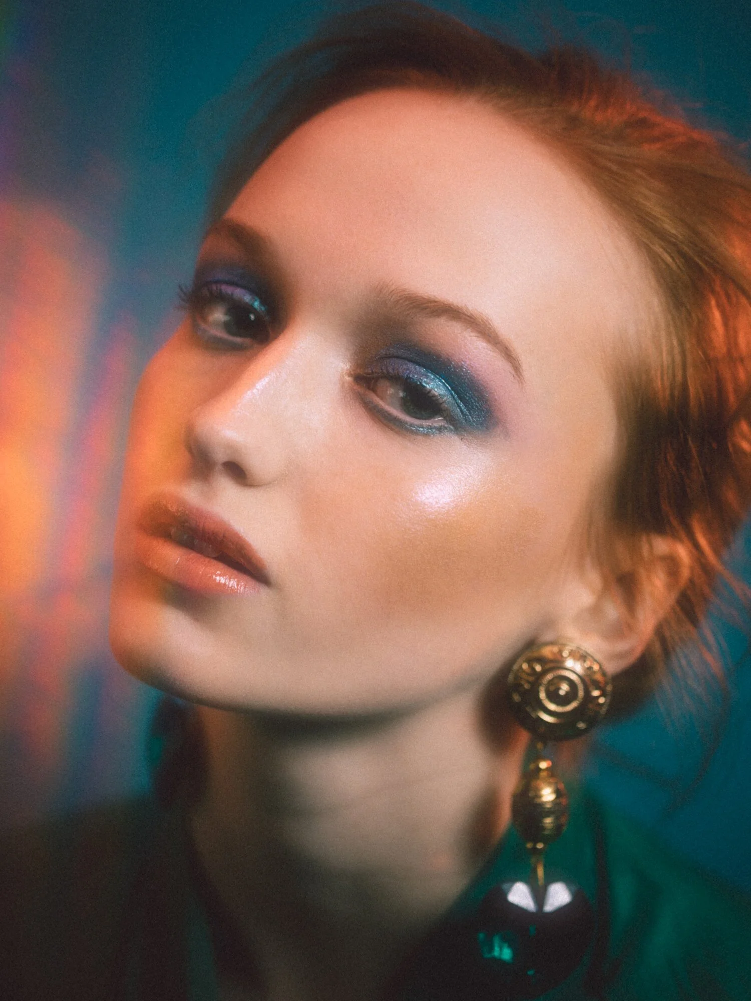 Close-up portrait of a woman with makeup featuring blue eyeshadow and glossy lips, wearing a large gold earring.