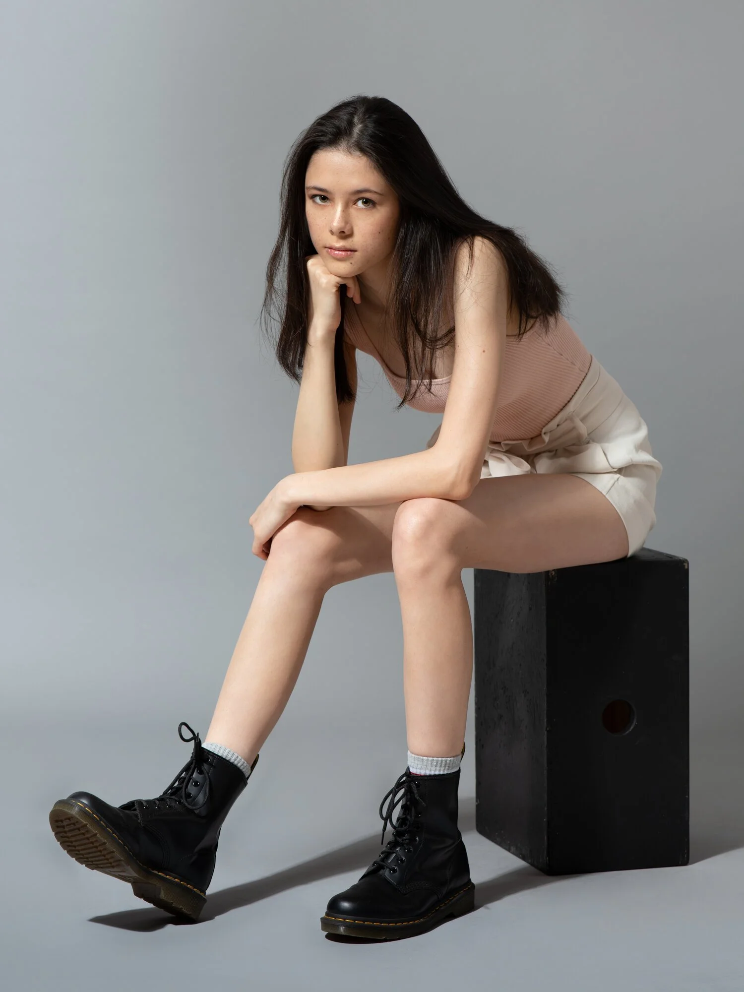 A young woman with long dark hair, sitting on a black box in front of a plain grey background. She is wearing a sleeveless pink top, beige shorts, grey socks, and black combat boots. She rests her chin on her hand and looks at the camera with a serious expression.