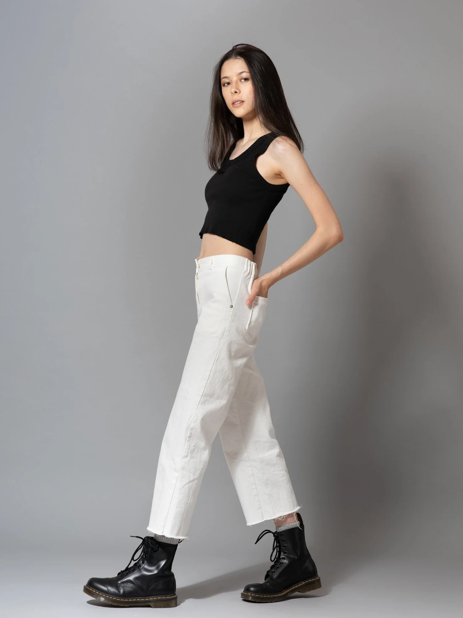 A young woman with long dark hair, wearing a black sleeveless cropped top, white high-waisted pants, and black lace-up boots, standing against a plain gray background with a slight sideways pose.