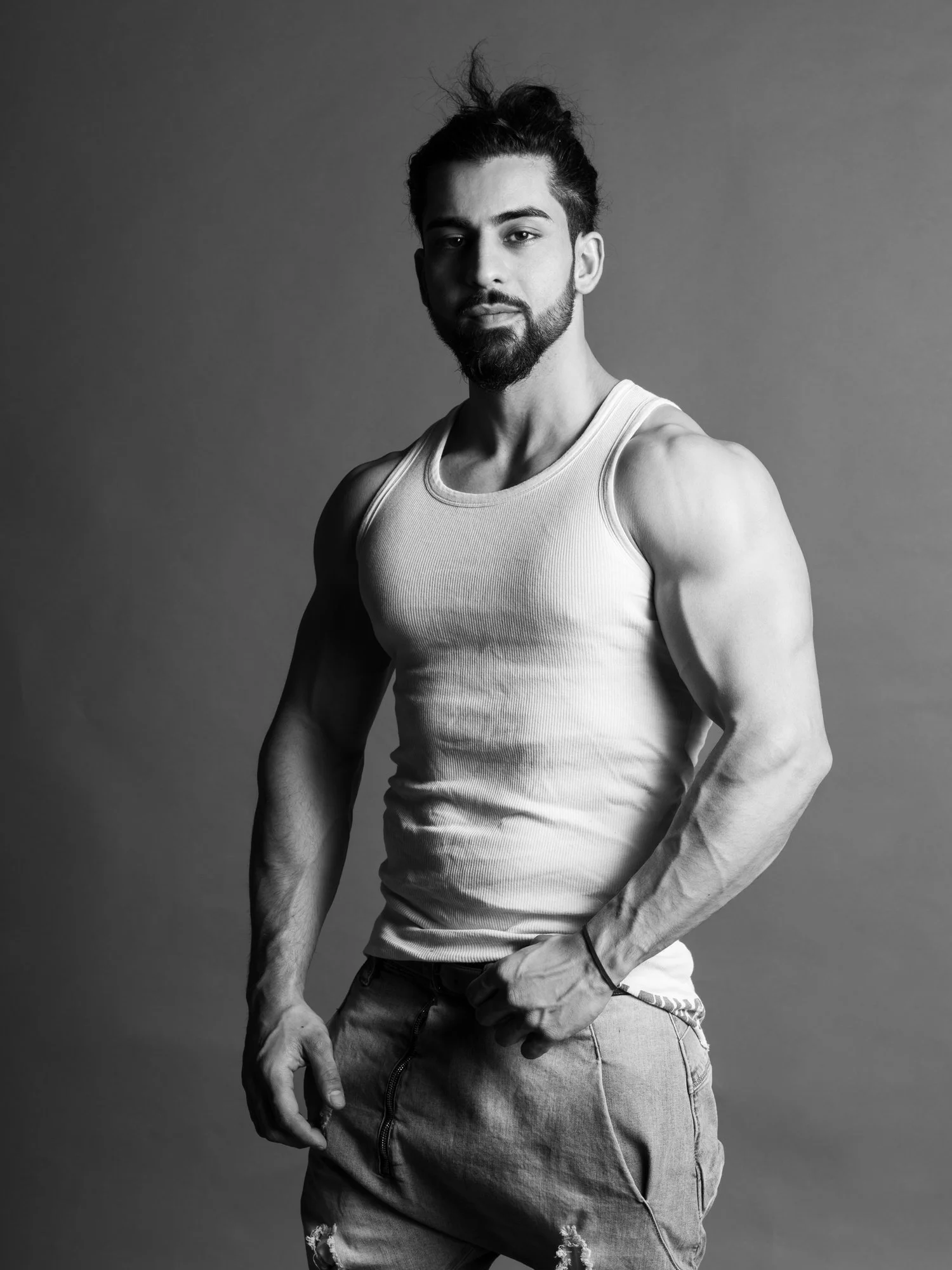 Black and white photo of a young man with dark hair, beard, wearing a tank top and ripped jeans, standing against a plain background.