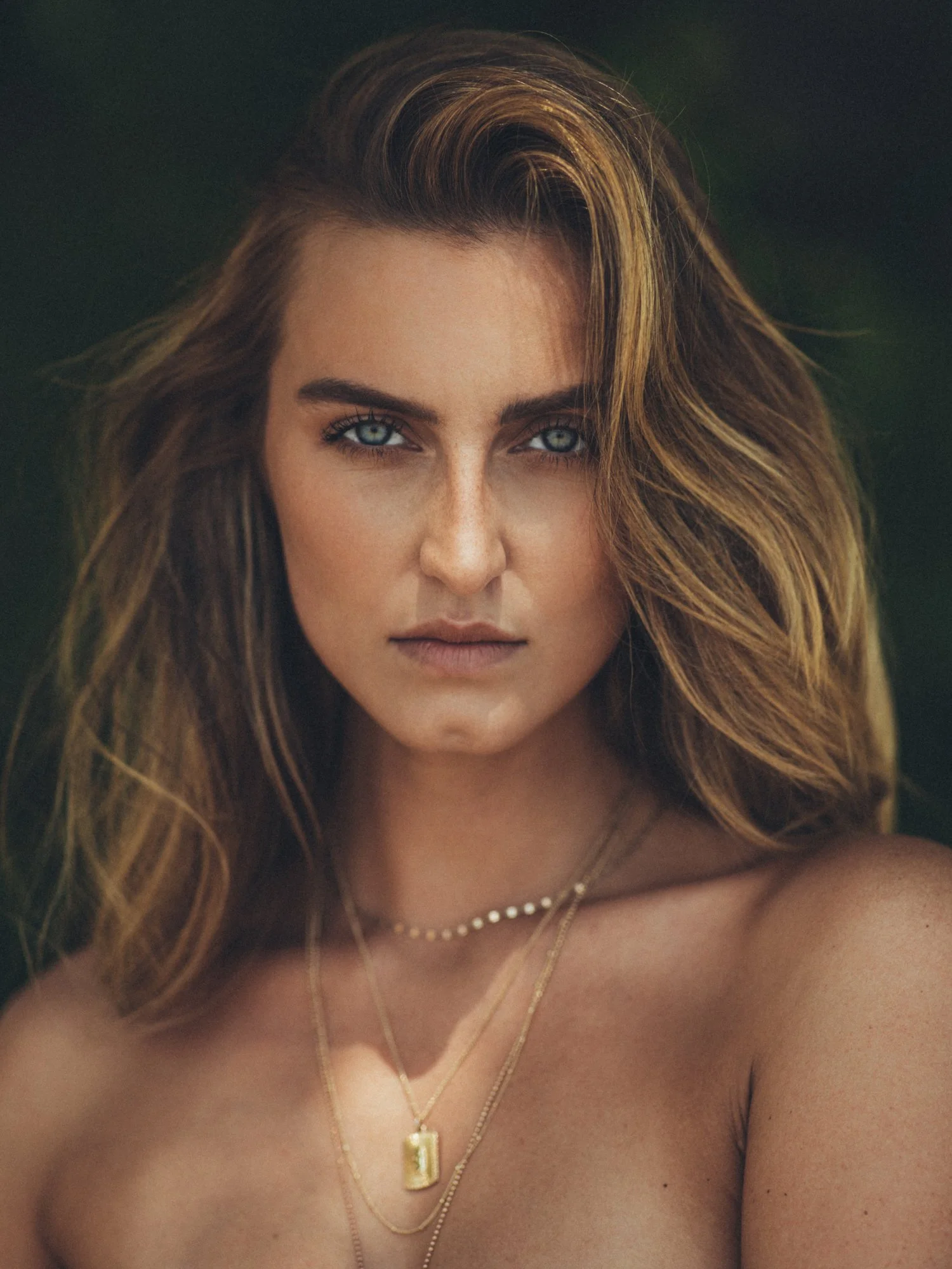 A woman with shoulder-length wavy brown hair and blue eyes looking directly at the camera, wearing layered gold necklaces, including one with a rectangular pendant, against a blurred green background.