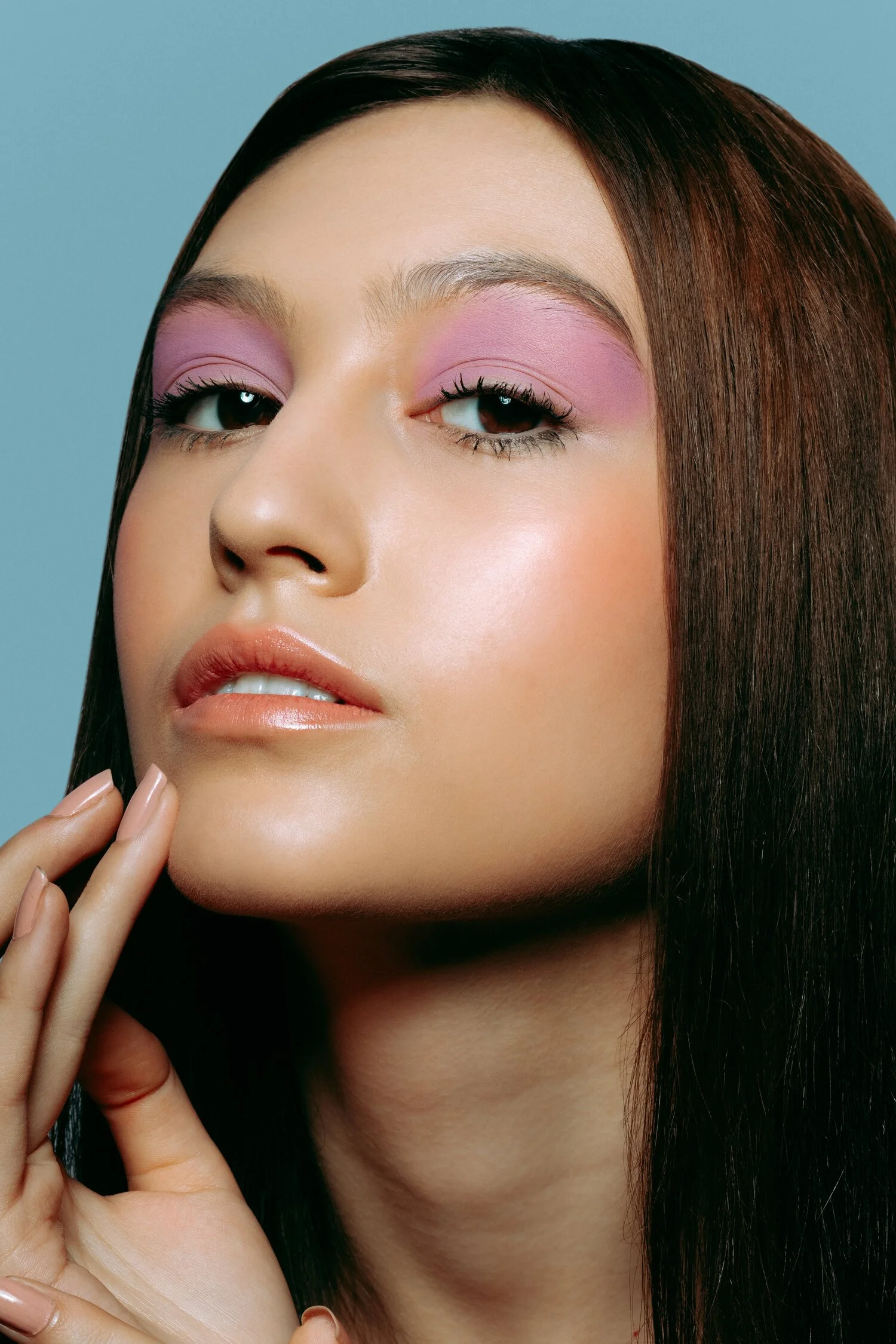Close-up of a woman with straight brown hair, pink eyeshadow, natural lipstick, and clear skin, against a light blue background.