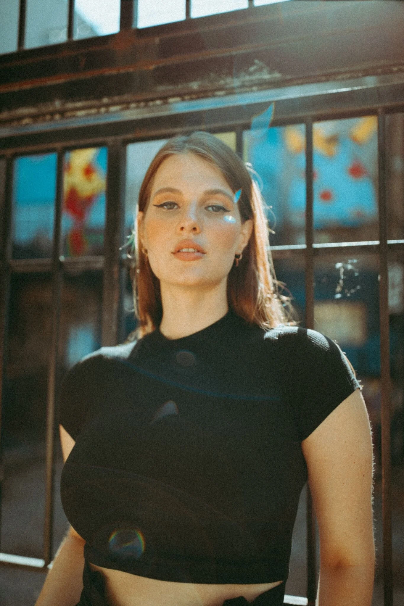 A young woman with shoulder-length brown hair and light makeup standing outdoors in front of a metal fence with colorful murals in the background, illuminated by sunlight.