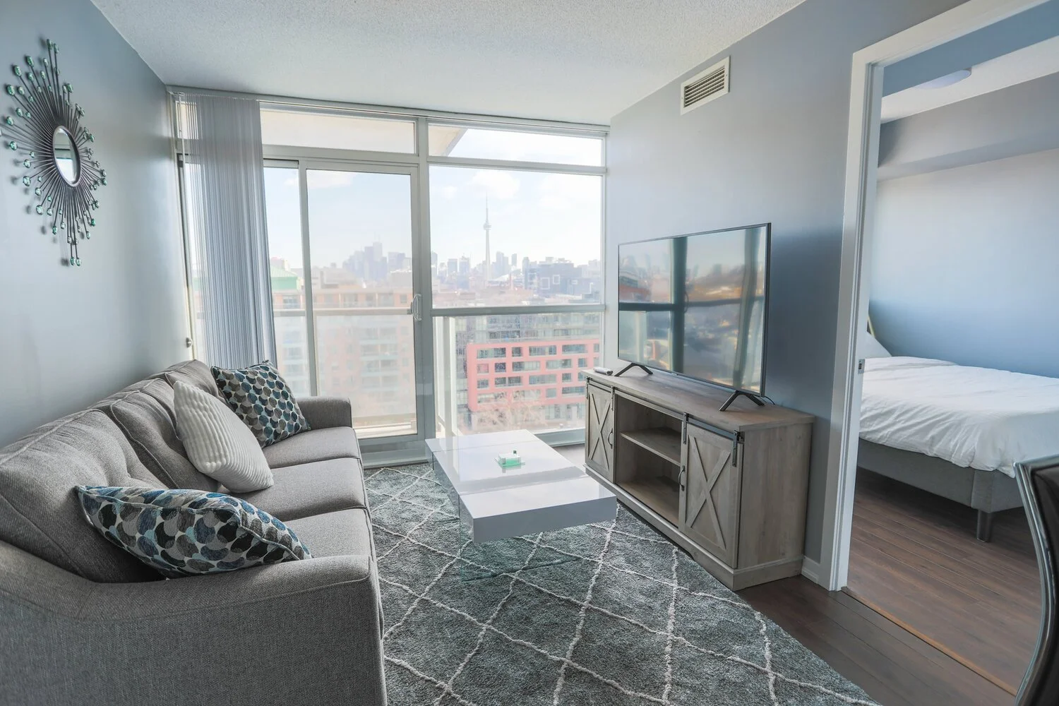 Living room with a gray sofa, patterned pillows, a white coffee table, a TV on a wooden stand, large windows showing a city skyline with a tower, and a glimpse of a bedroom with a bed.