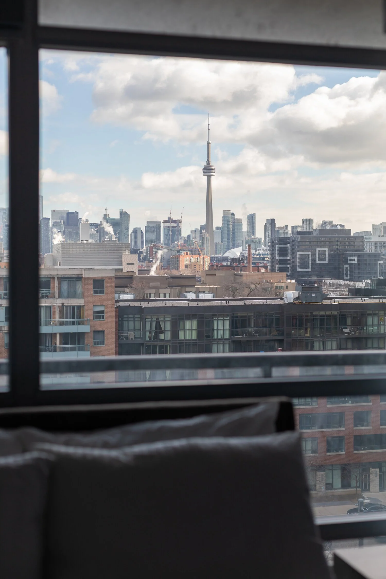 View of Toronto skyline through a window, featuring the CN Tower and surrounding skyscrapers, with a partially visible couch in the foreground.