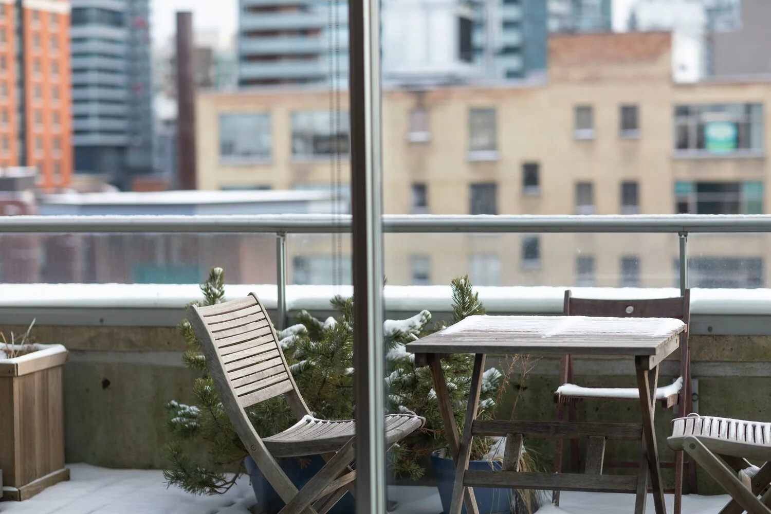 A snowy balcony with a wooden table and chairs, small pine trees in pots, and a railing overlooking a cityscape with various buildings.