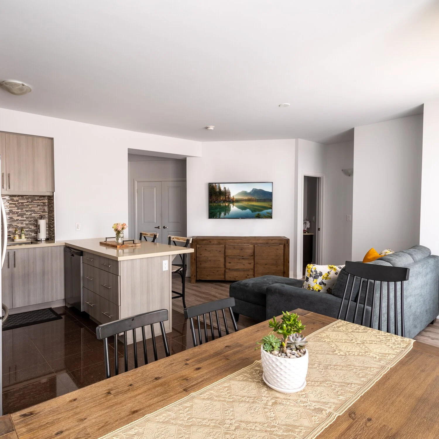 Open-concept living room and kitchen with dining table, gray sofa, flat-screen TV, cabinets, and decor.