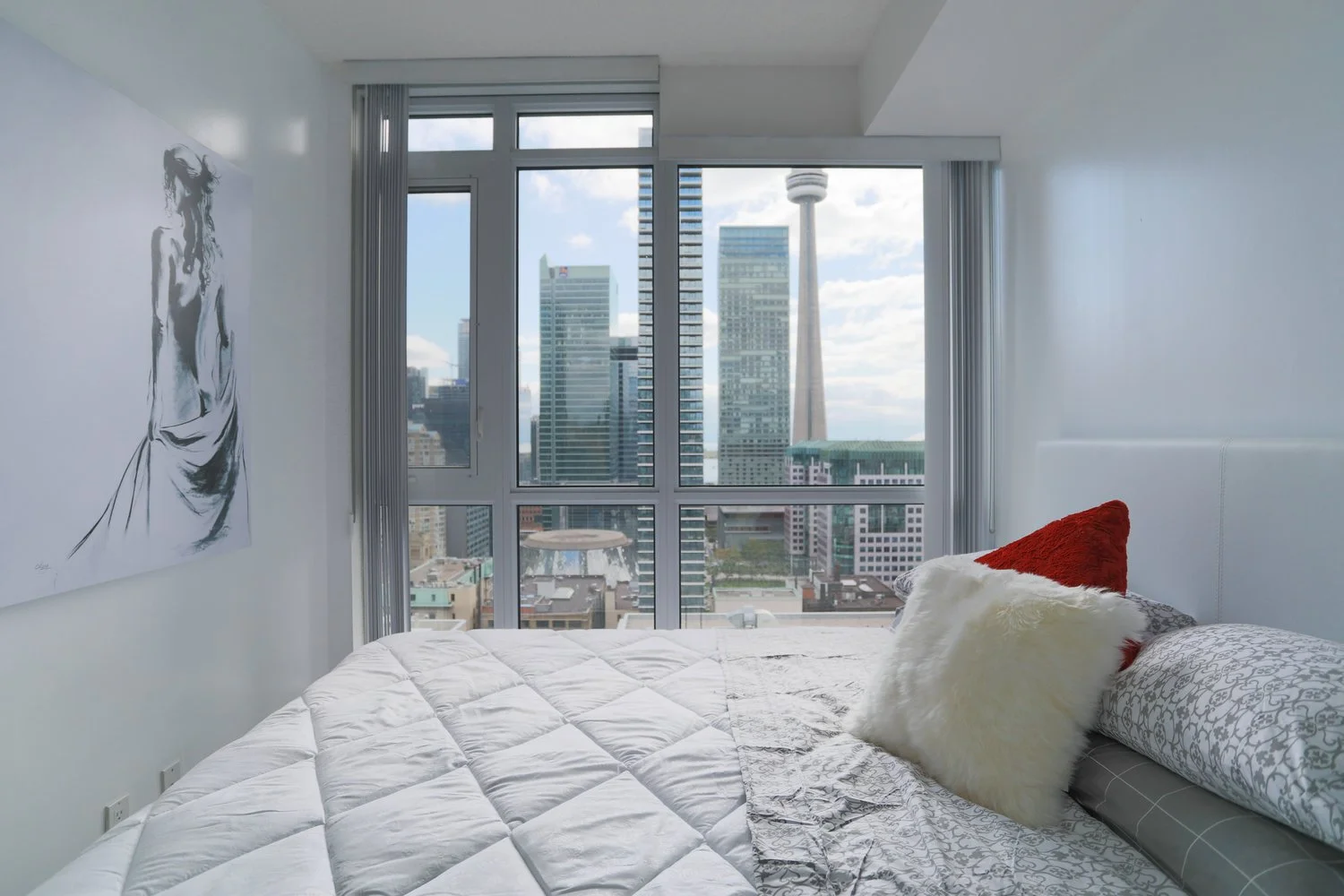 Modern bedroom with a large window showing city skyscrapers and the CN Tower in the background. The bed has white and patterned bedding with red and white fluffy pillows. There is a black and white artwork of a woman on the wall.