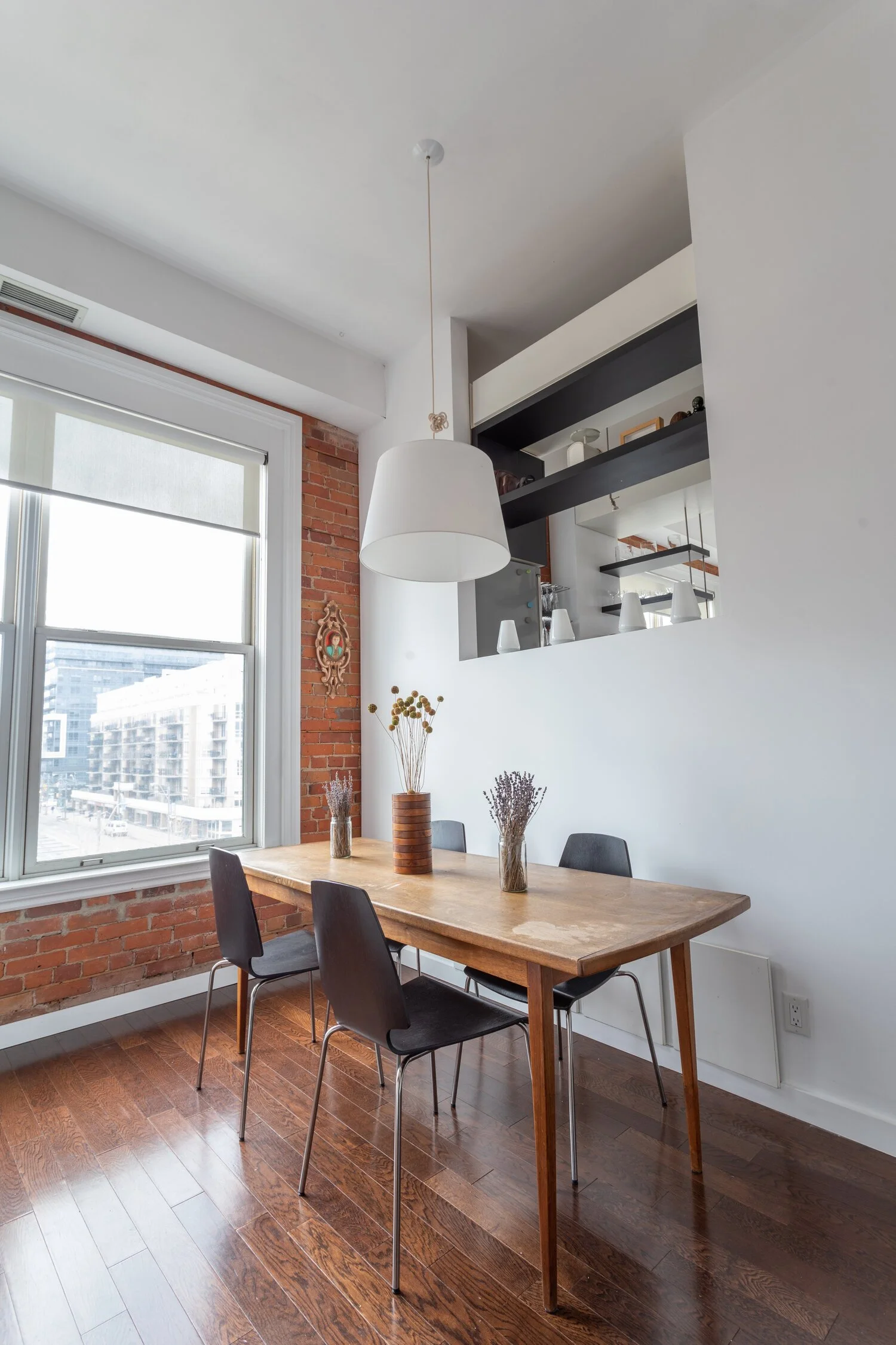 A dining area in a modern apartment with a wooden table, four black chairs, vases with dried flowers, a large window with a city view, a brick wall, and a white hanging pendant light.