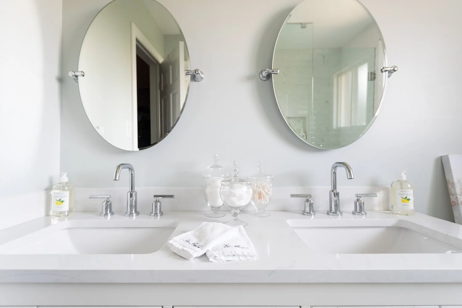Double bathroom vanity with two oval mirrors, chrome faucets, soap dispensers, glass jars with cotton and cotton swabs, and white towels on a white countertop.