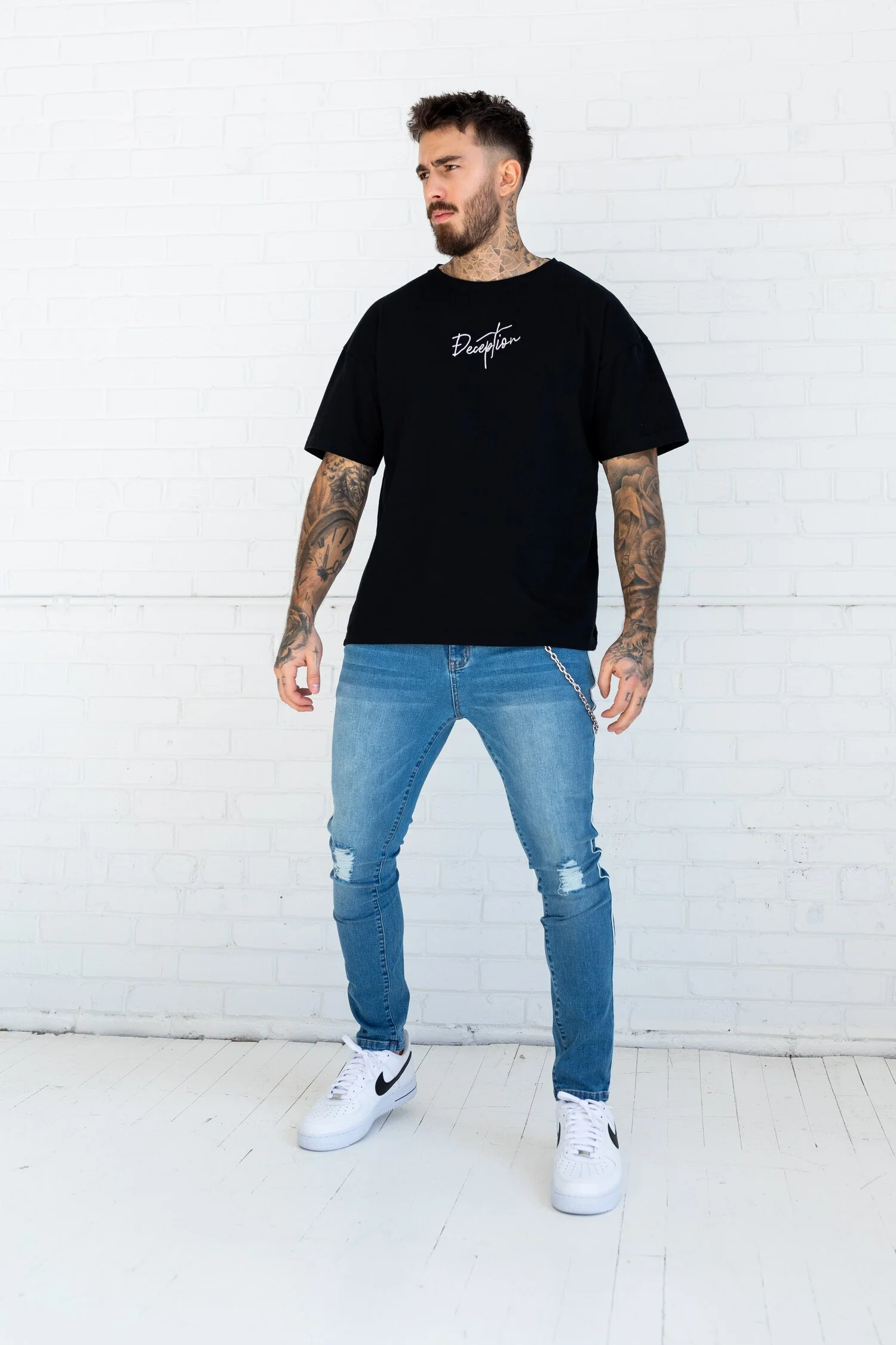 Young man with tattoos posing against a white brick wall in a black Deception t-shirt.