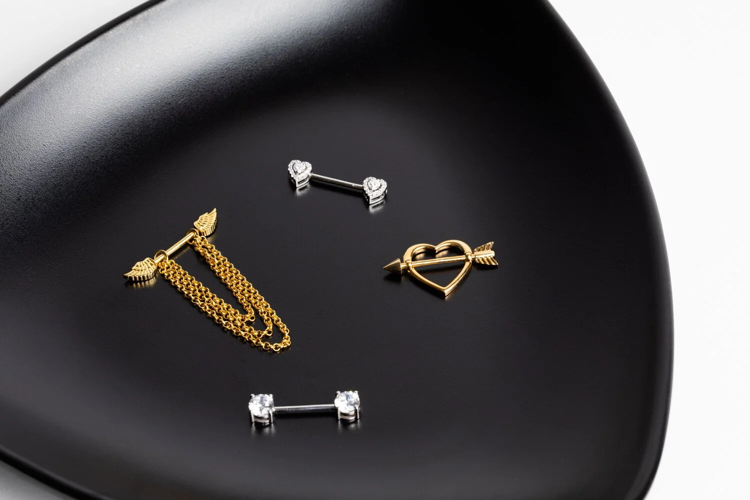 Gold and silver nose jewelry shaped like hearts and arrows on a black plate.